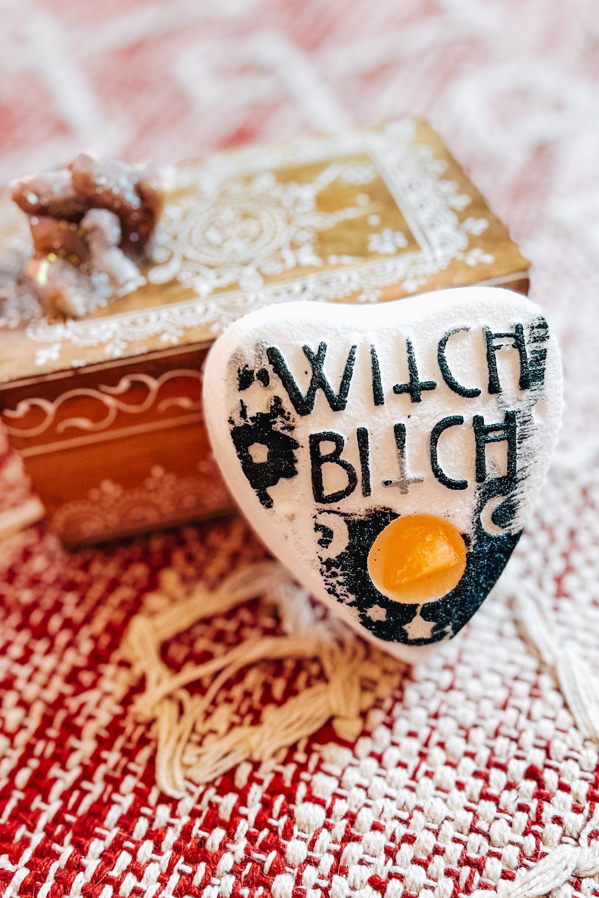 White crystal bath bomb with black lettering that says “witch bitch” and citrine crystal center resting on woven red and white fabric beside decorative wooden box