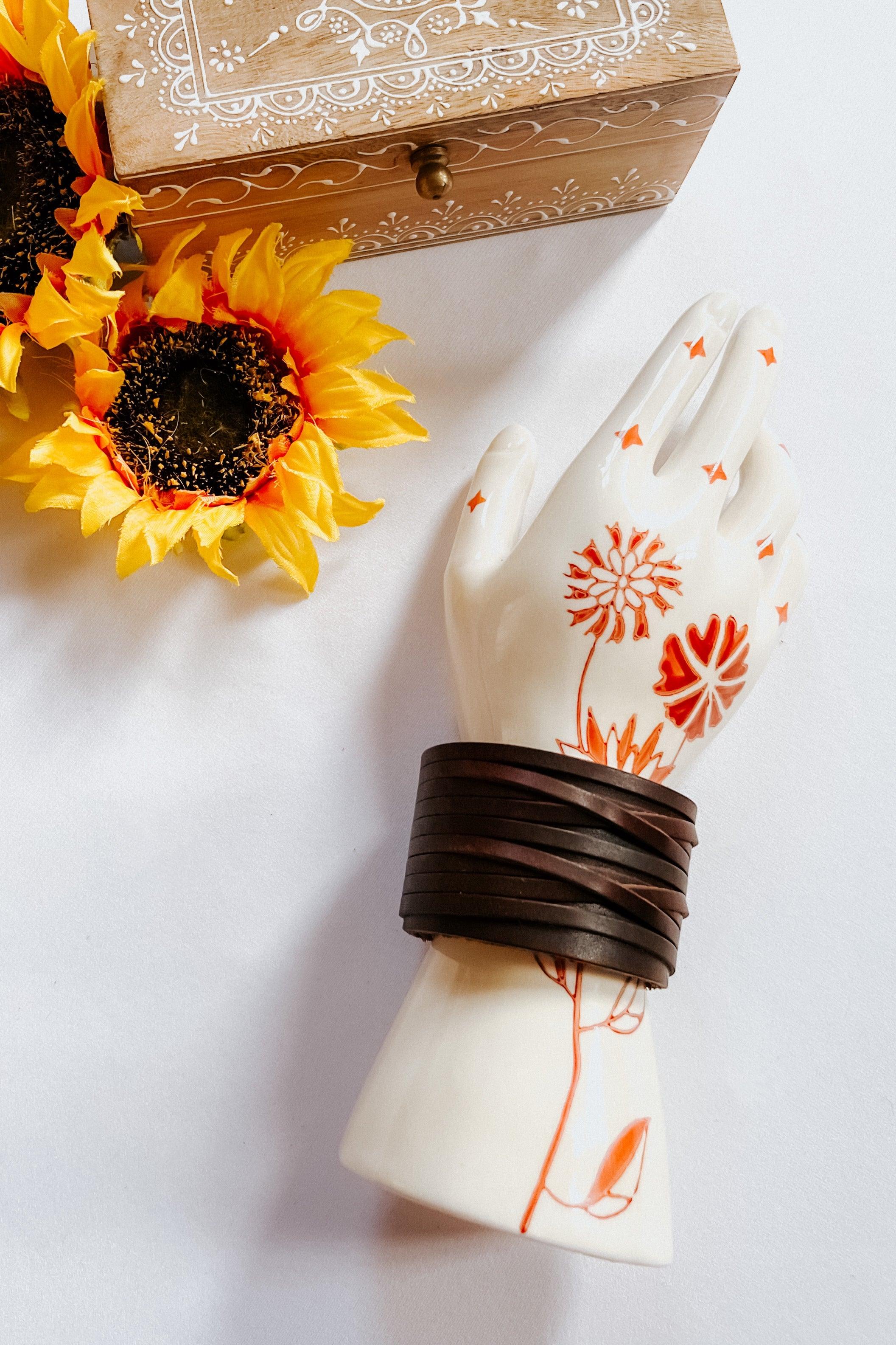 Brown leather cuff bracelet wrapped around ceramic hand with orange floral design beside wooden box and sunflowers