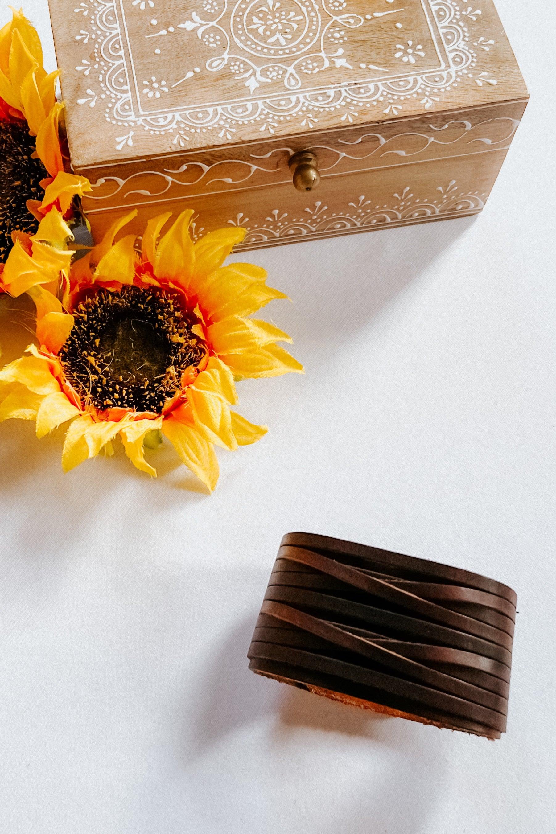 Dark brown leather cuff bracelet with layered design next to wooden box and bright artificial sunflowers on white surface