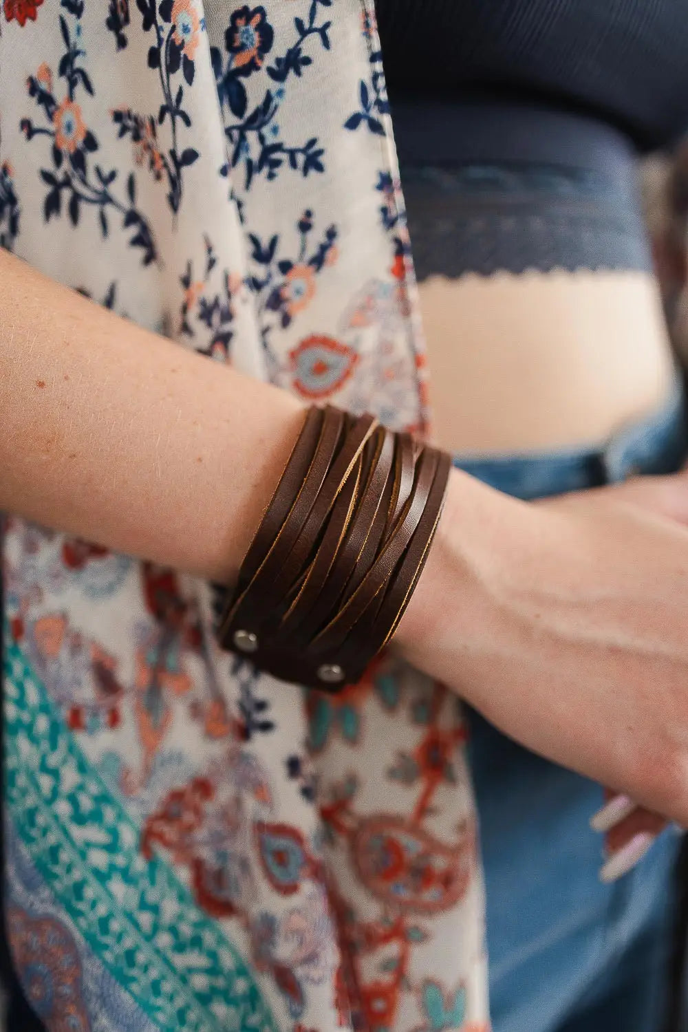 Brown leather cuff bracelet with layered straps worn on wrist against floral patterned fabric and casual outfit