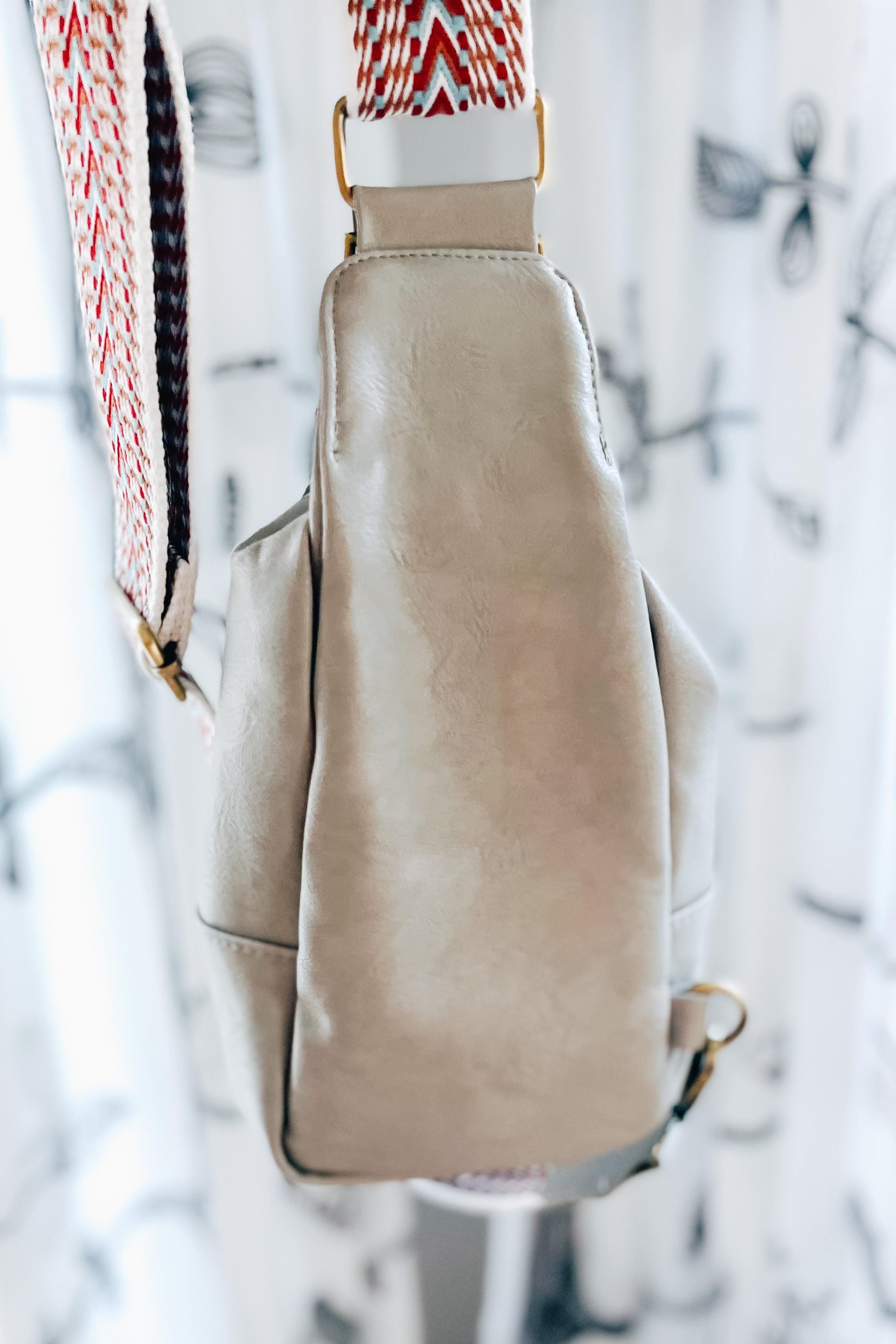 Side view of a grey vegan leather sling bag with bold patterned strap hanging indoors against patterned curtains