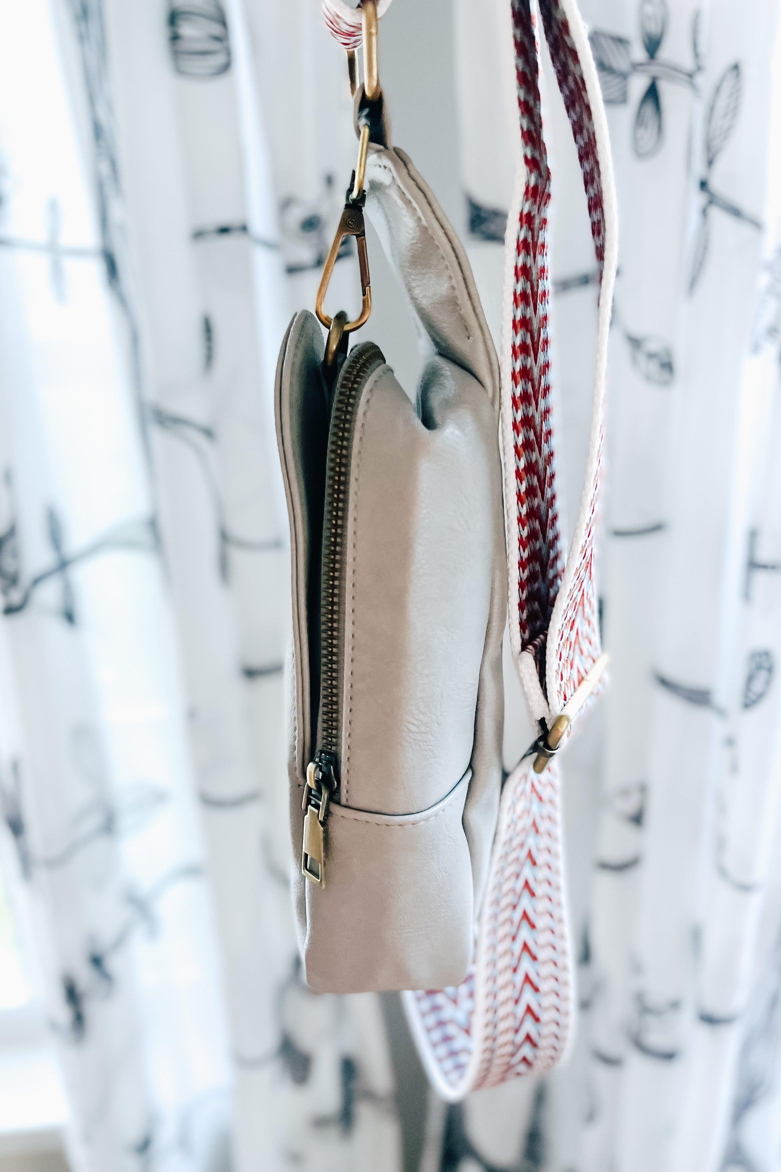 Small grey vegan leather sling bag with geometric patterned adjustable strap hanging indoors by window with floral curtains
