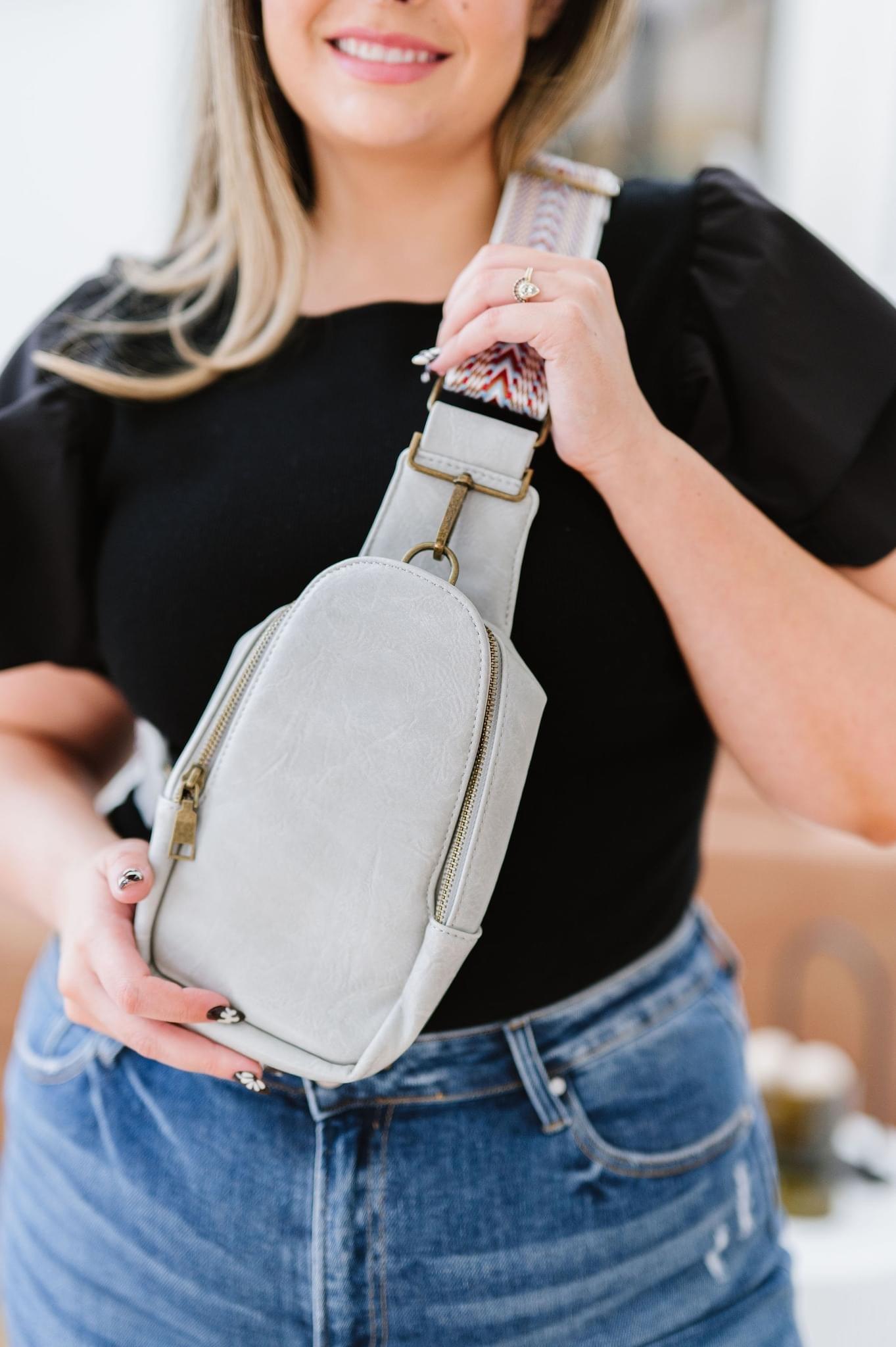 Woman holding gray vegan leather sling bag with patterned strap, wearing black top and blue denim jeans indoors
