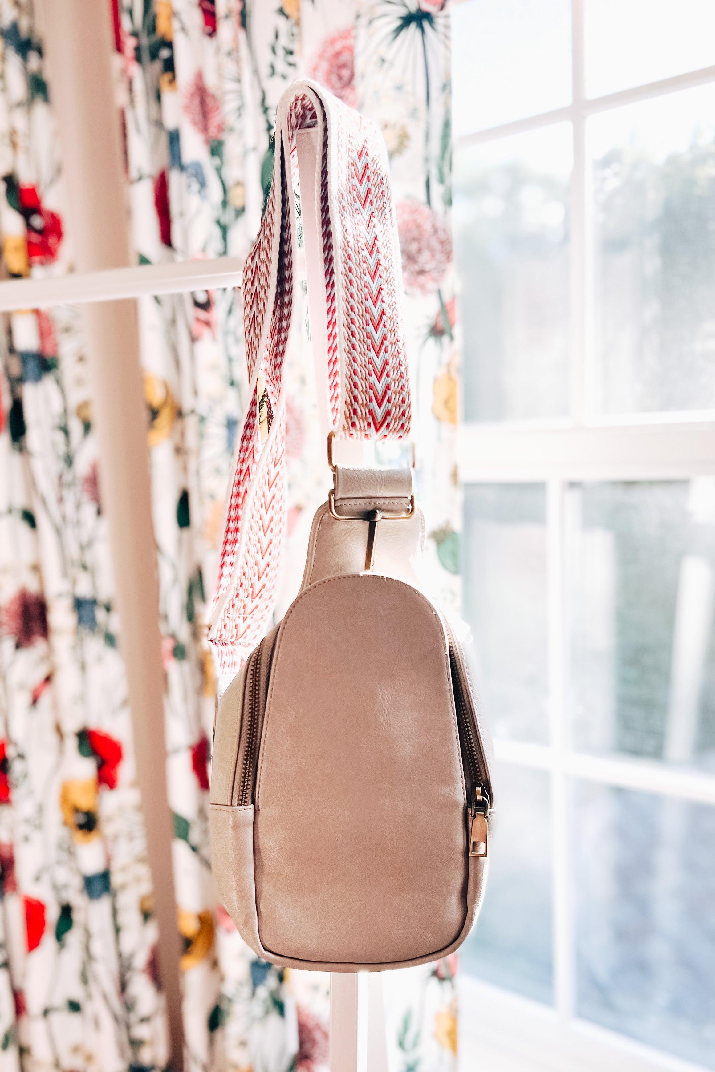 Grey vegan leather sling bag with patterned fabric strap hanging indoors near floral curtains and window light