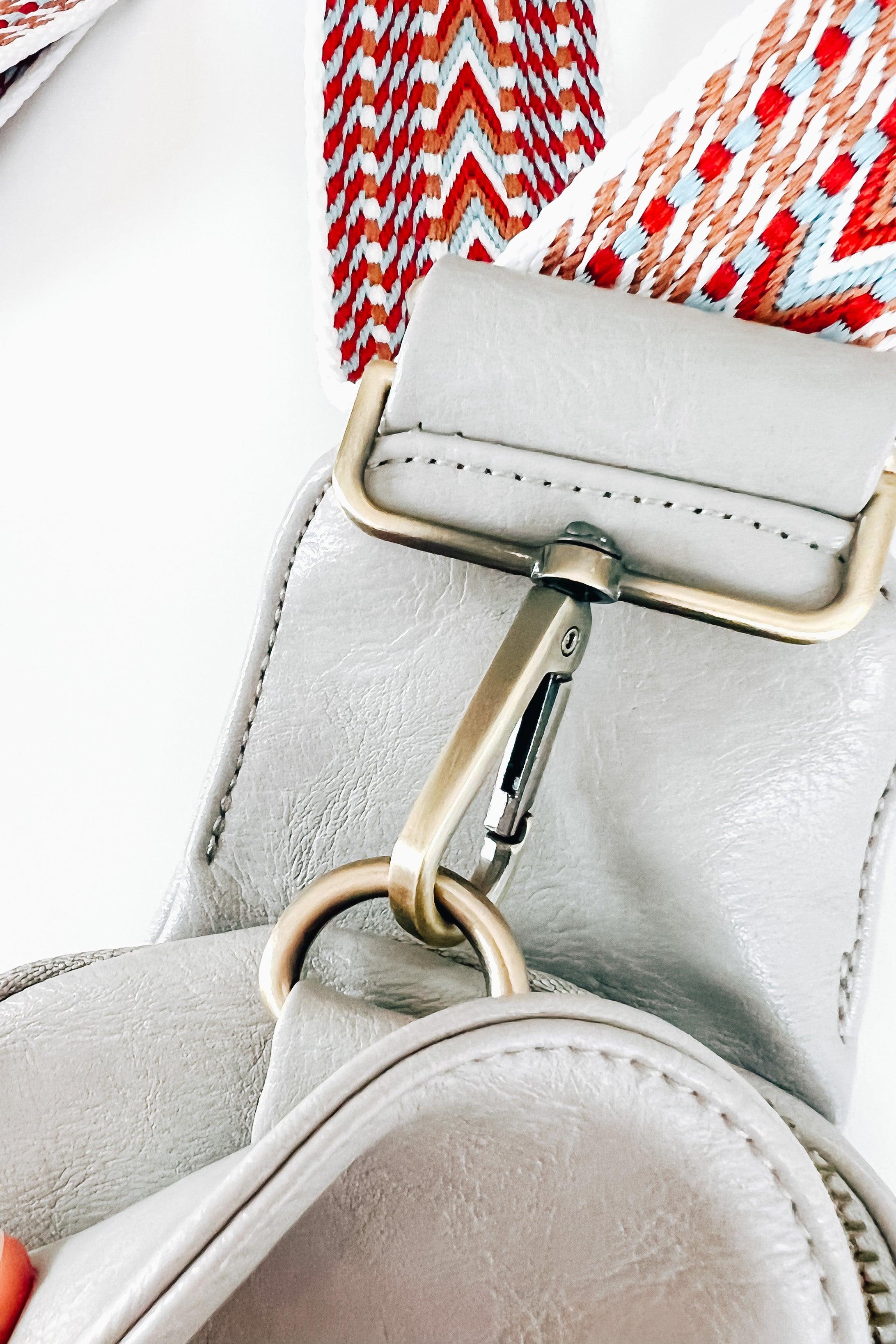 Close-up of grey vegan leather sling bag attaching to strap with patterned red and white woven fabric and brass hardware