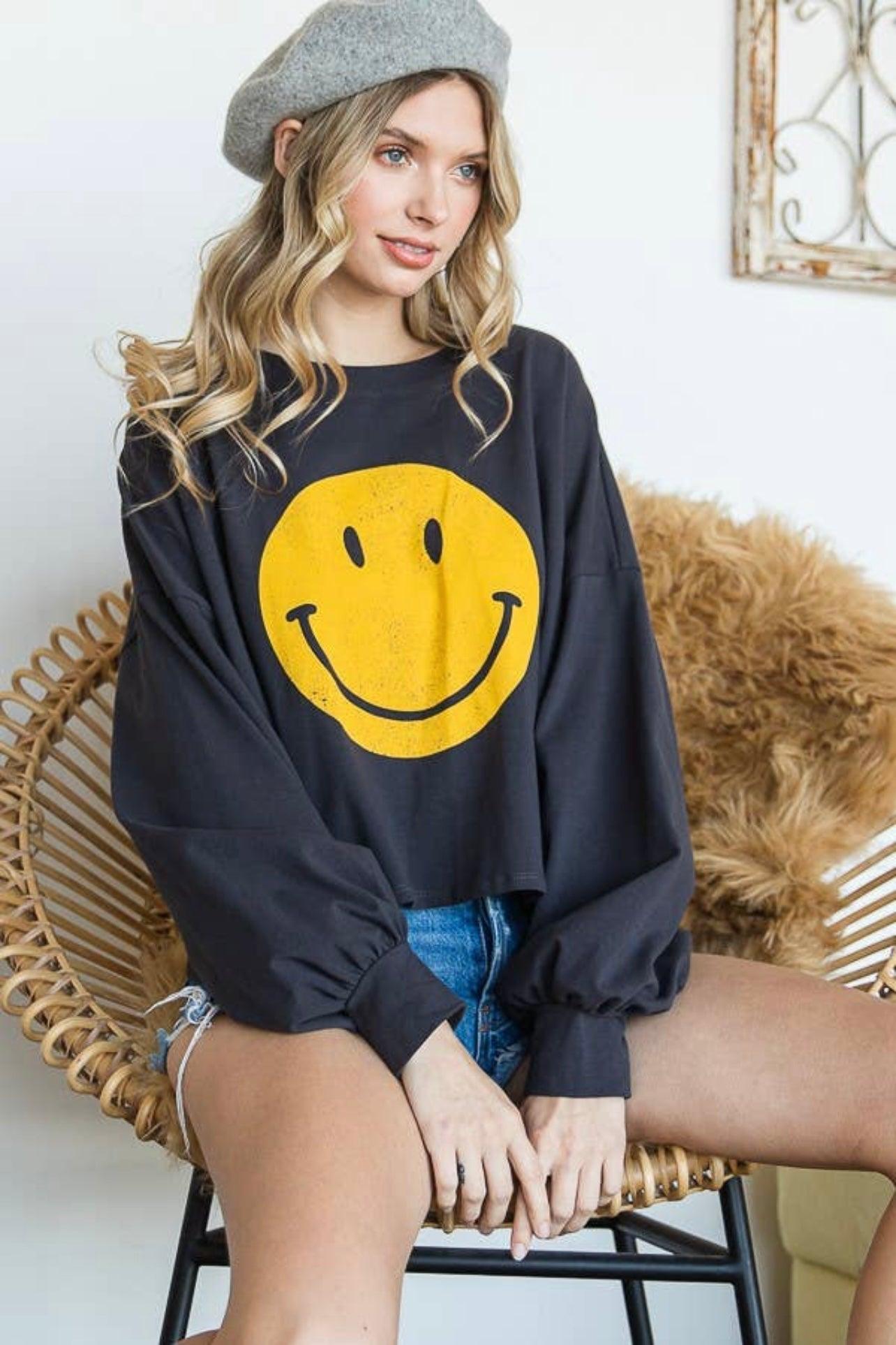Woman wearing charcoal long sleeve crop top with large yellow smiley face sitting in wicker chair indoors