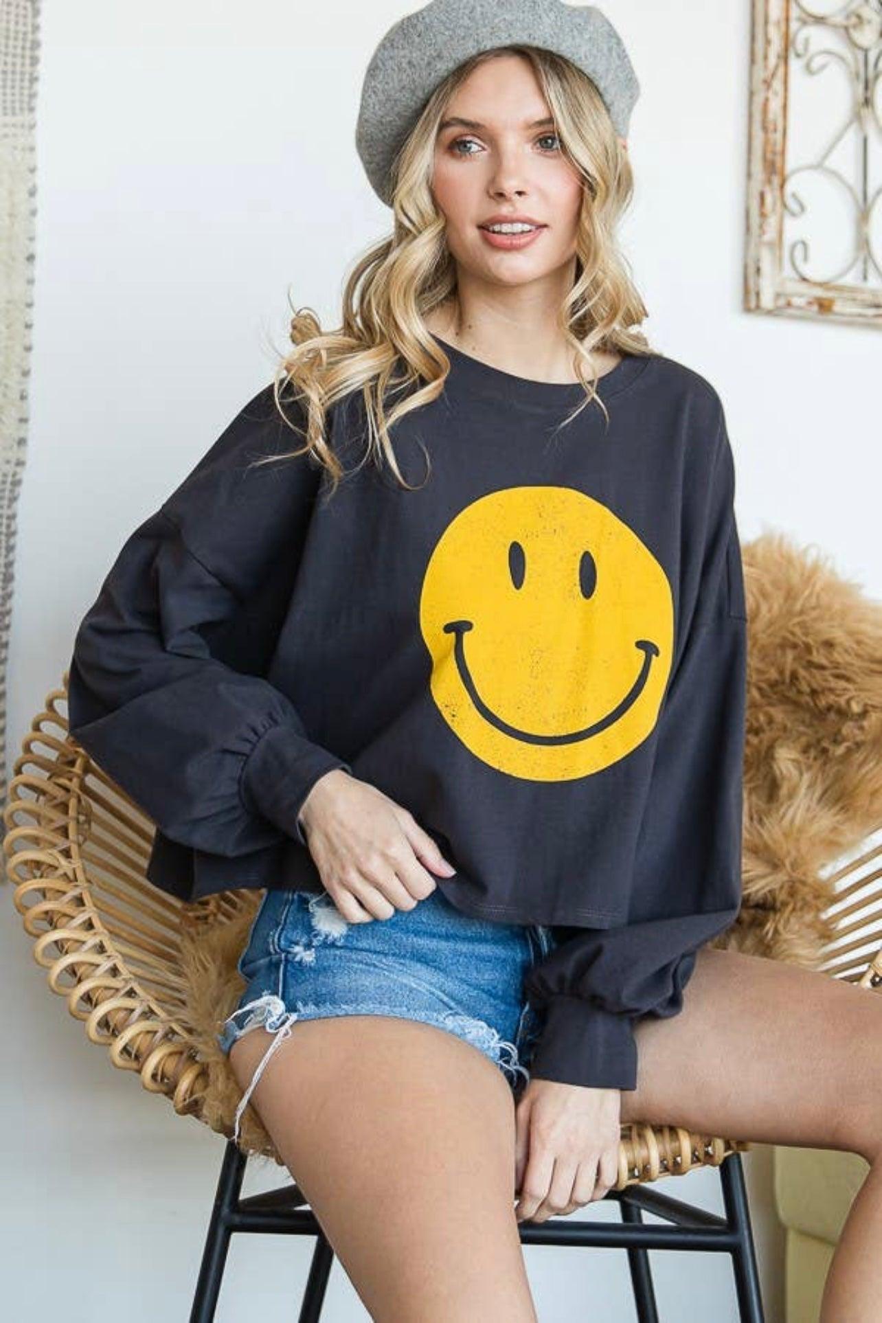 Charcoal long sleeve crop top with yellow smiley face worn with denim shorts and grey beret seated on rattan chair
