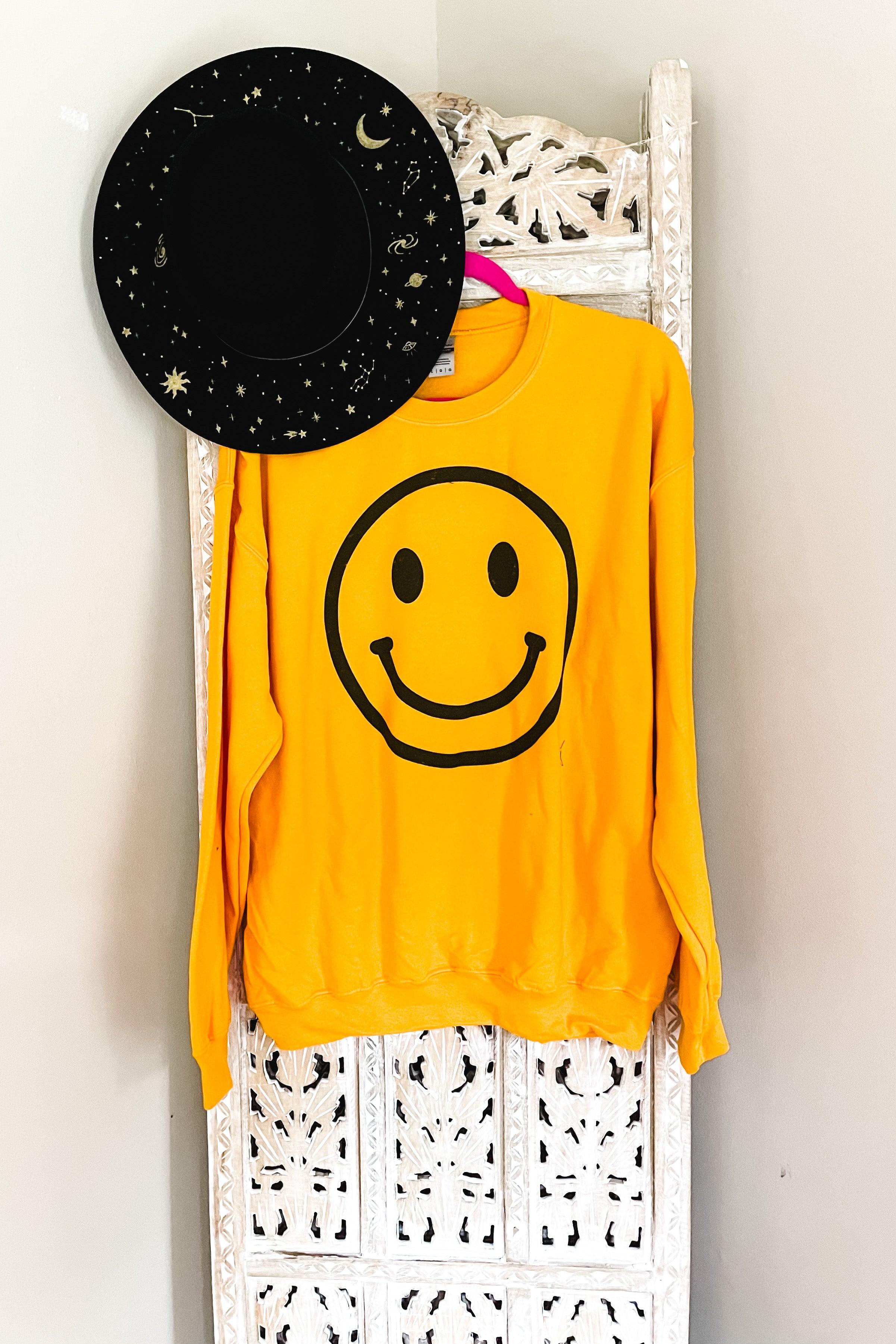 Yellow sweatshirt with oversized black smiley face graphic hanging on carved white wooden screen with black starry hat