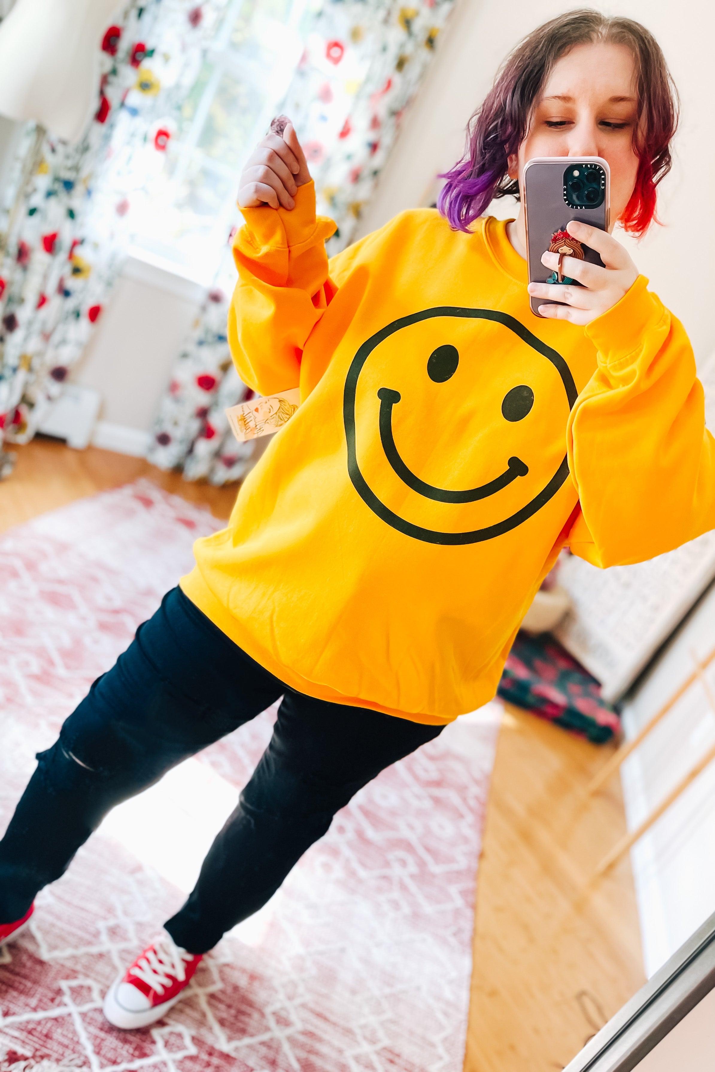 Woman with multi-colored hair wearing bright yellow sweatshirt with large black smiley face taking mirror selfie indoors