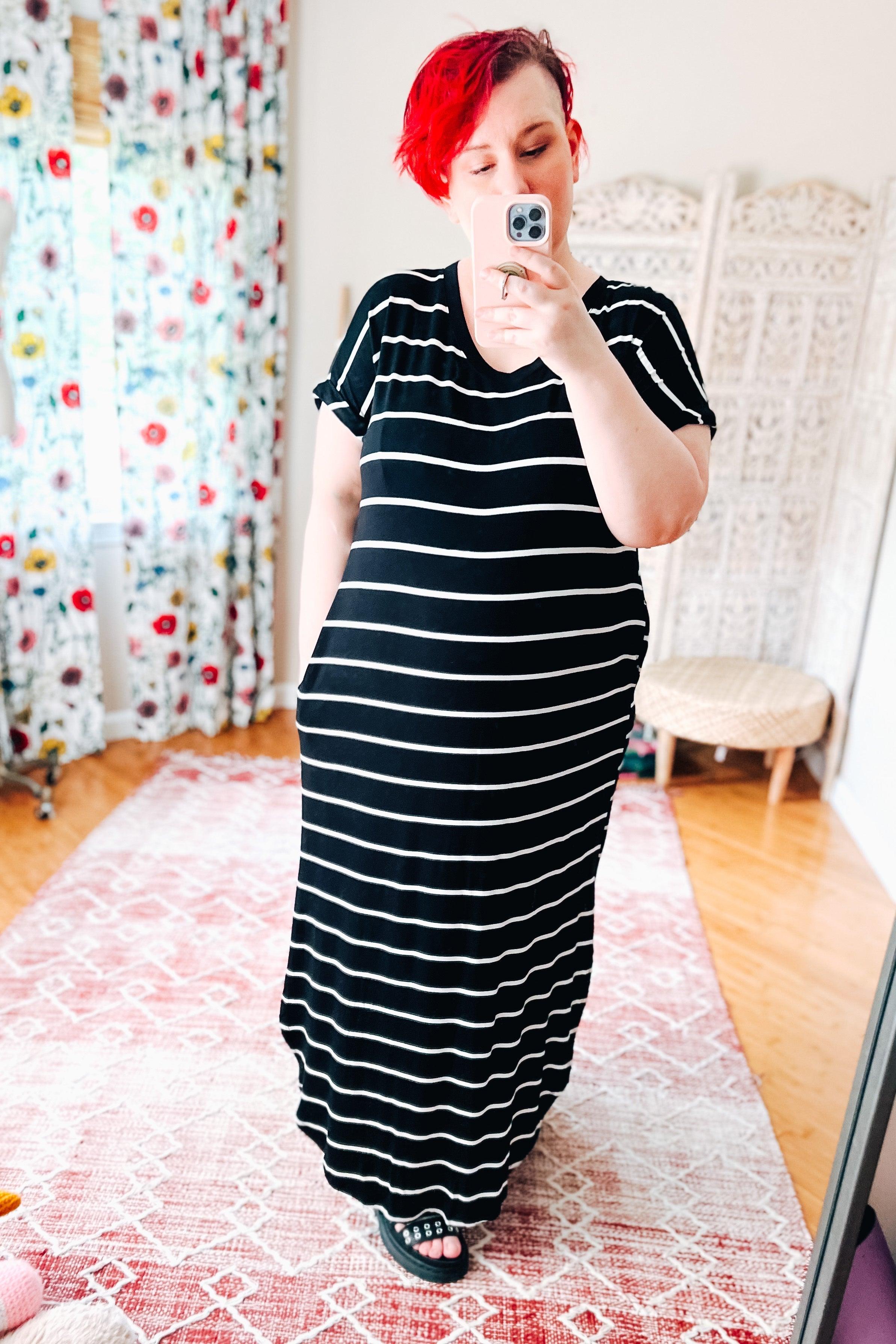 Woman with red hair wearing a black and white striped maxi dress taking a mirror selfie in a bright room