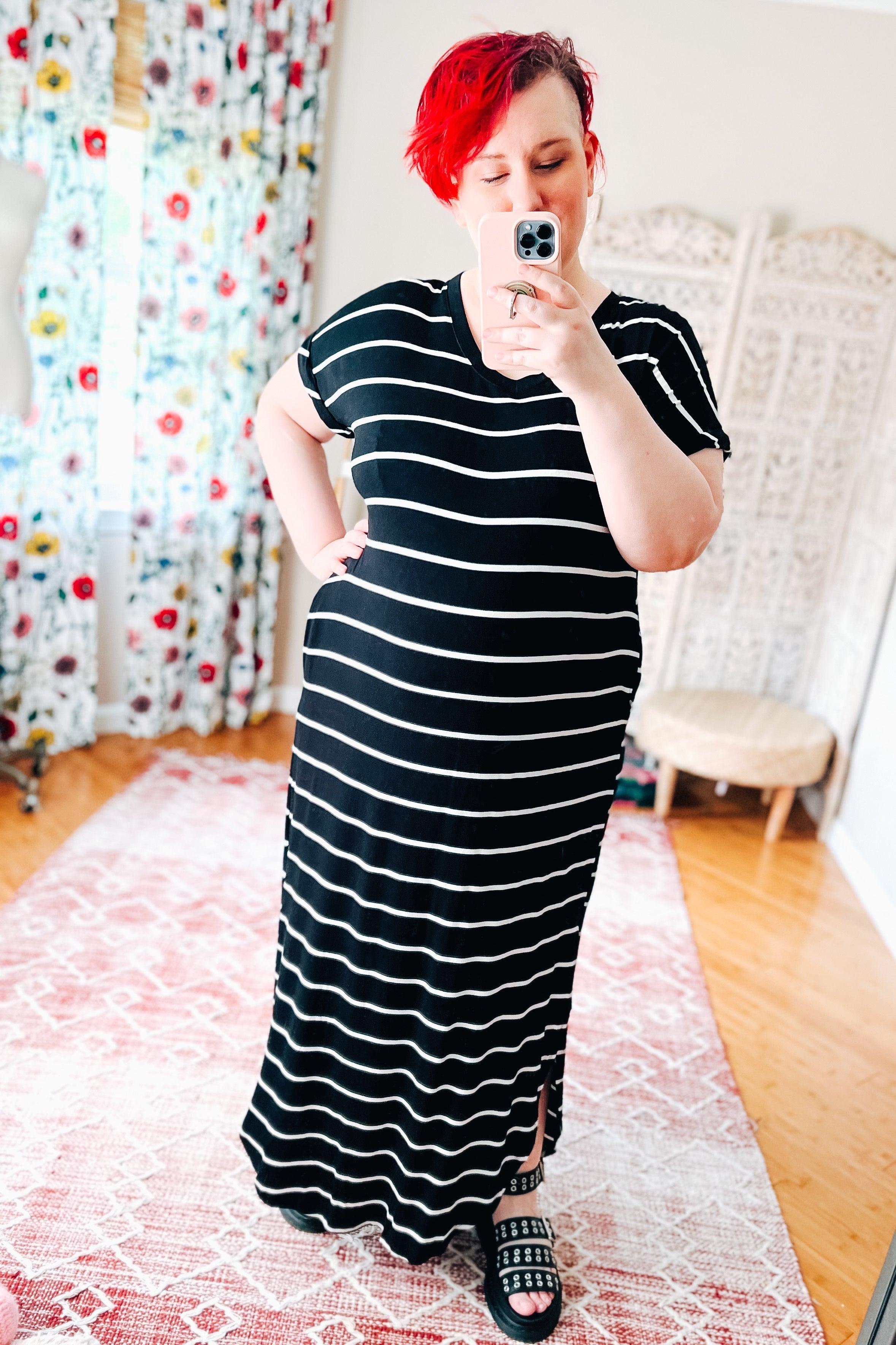 Woman with red hair wearing a black and white striped maxi dress and studded sandals taking a mirror selfie indoors