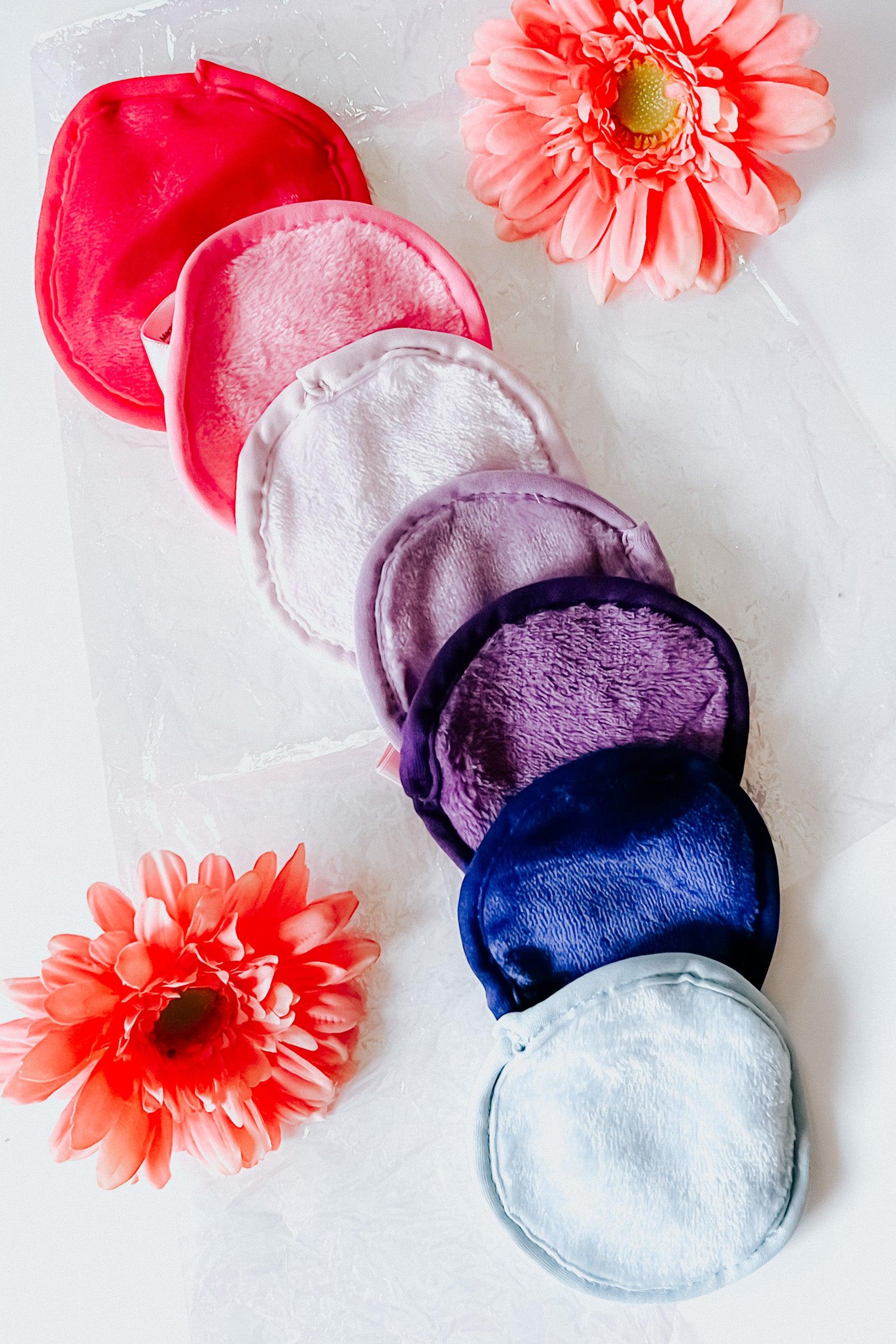 Seven plush reusable makeup remover pads in pink, purple, and blue shades with two coral flowers on white background