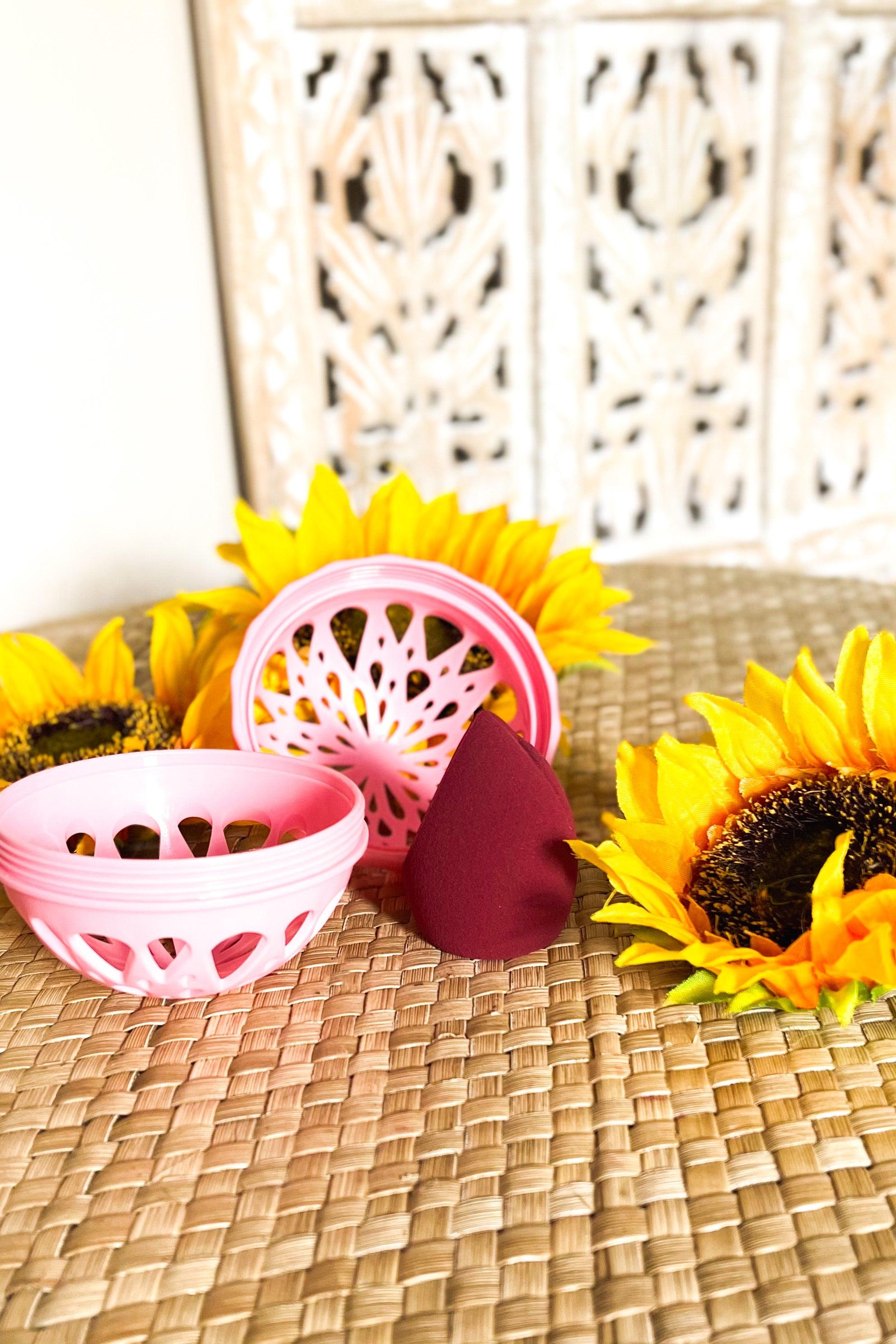 Pink vented container with a burgundy makeup sponge surrounded by bright sunflower blooms on woven surface