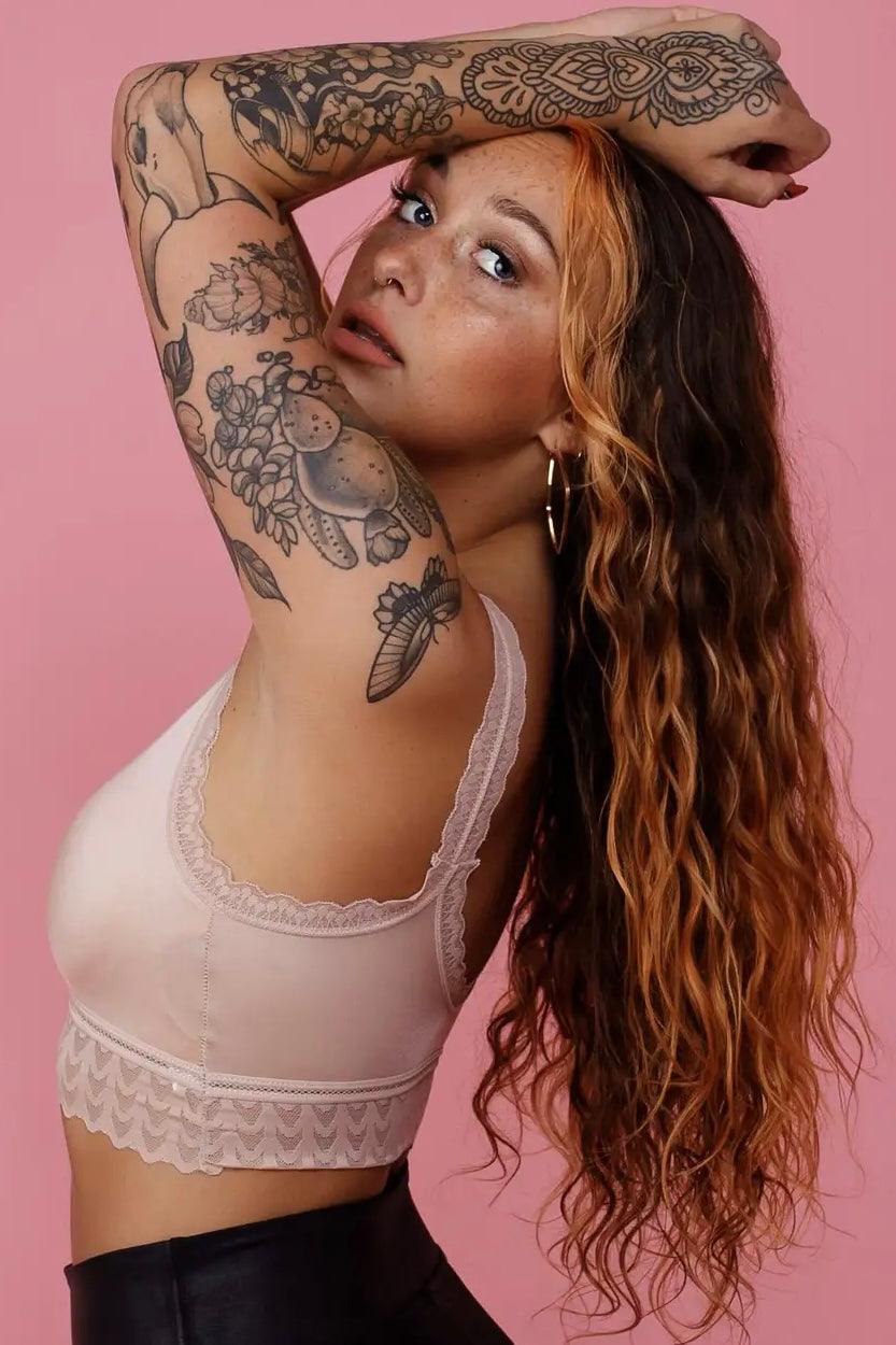 Woman with long wavy hair wearing a pink lace bralette and showcasing bold floral tattoos against a pink background
