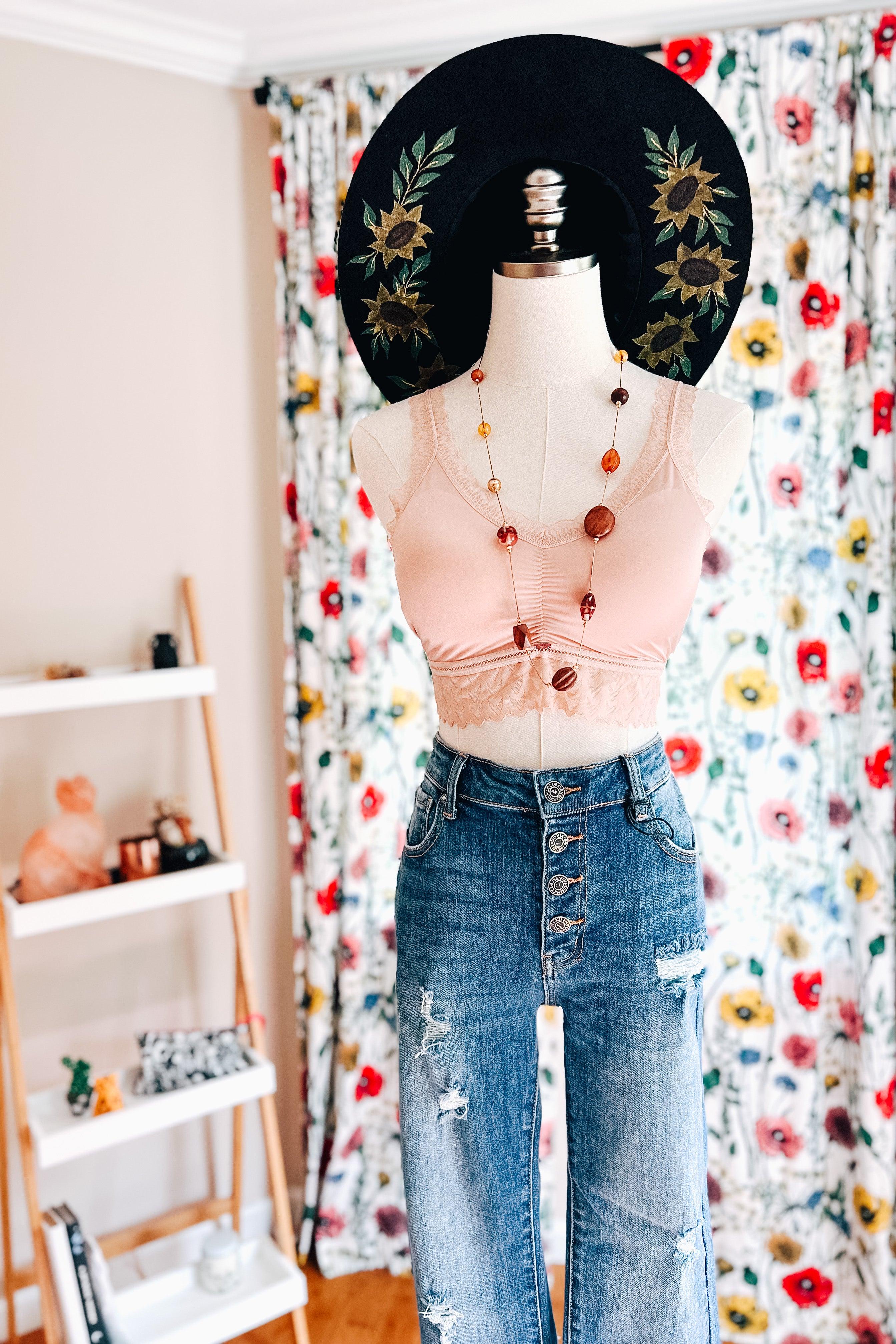 Mannequin wearing a soft pink bralette styled with beaded necklace, distressed high-waisted jeans, and a black floral hat indoors