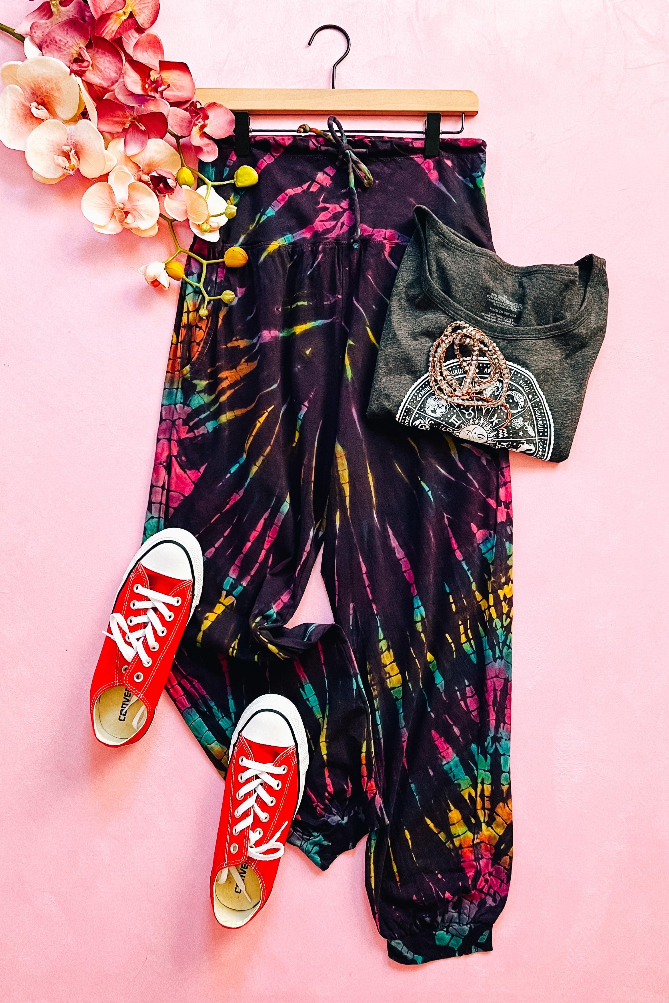 Purple Passion tie dye joggers styled with graphic tee, beaded necklace, and red canvas sneakers on pink surface
