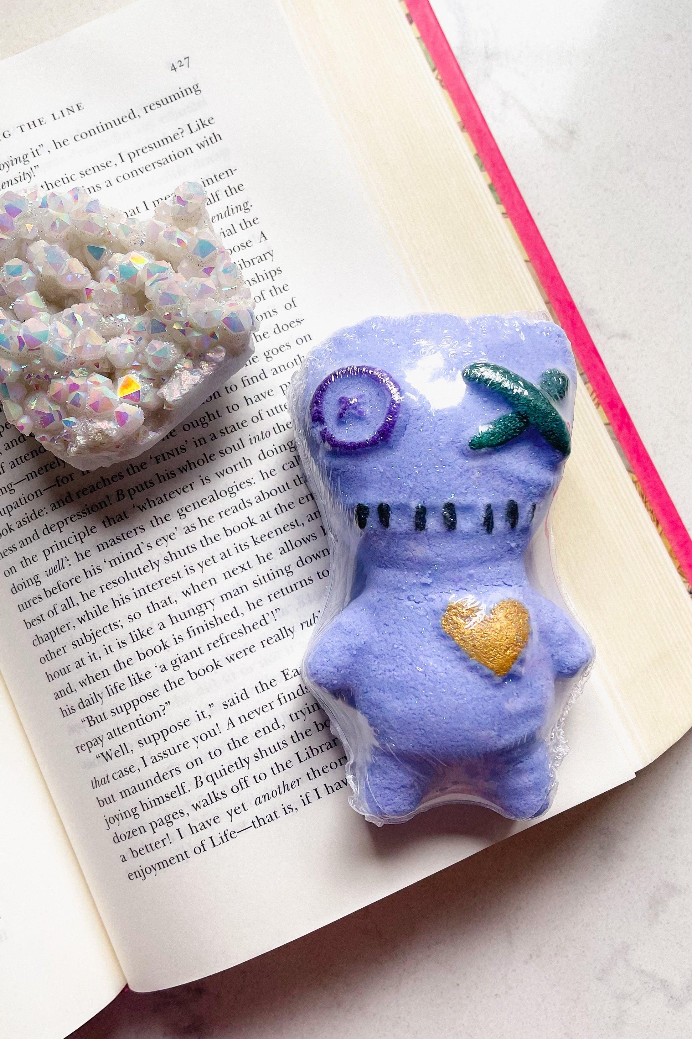 Purple bath bomb shaped like a voodoo doll with stitched details and a gold heart on an open book page