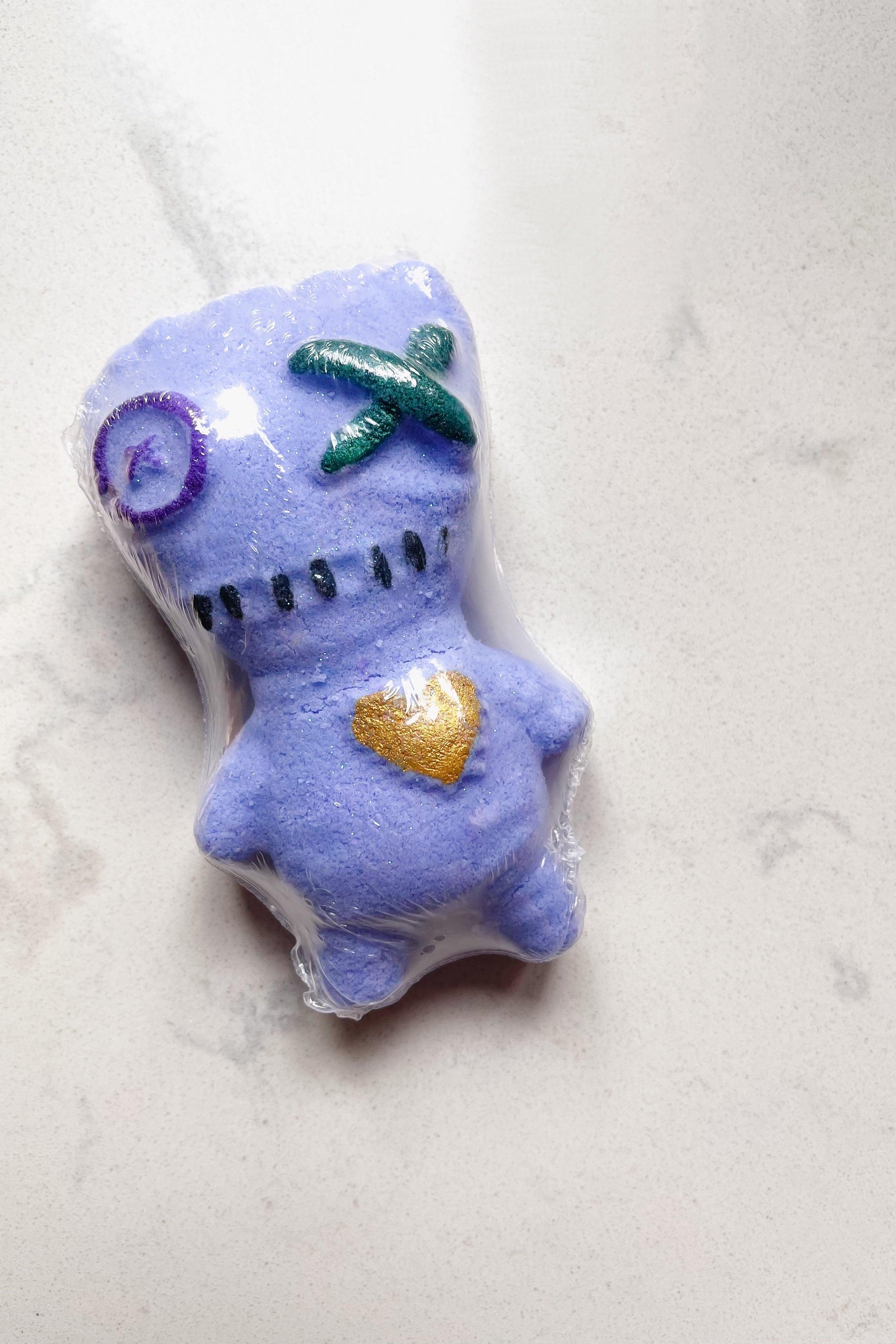 Purple voodoo doll shaped bath bomb with stitched facial features and gold heart on torso wrapped in clear plastic