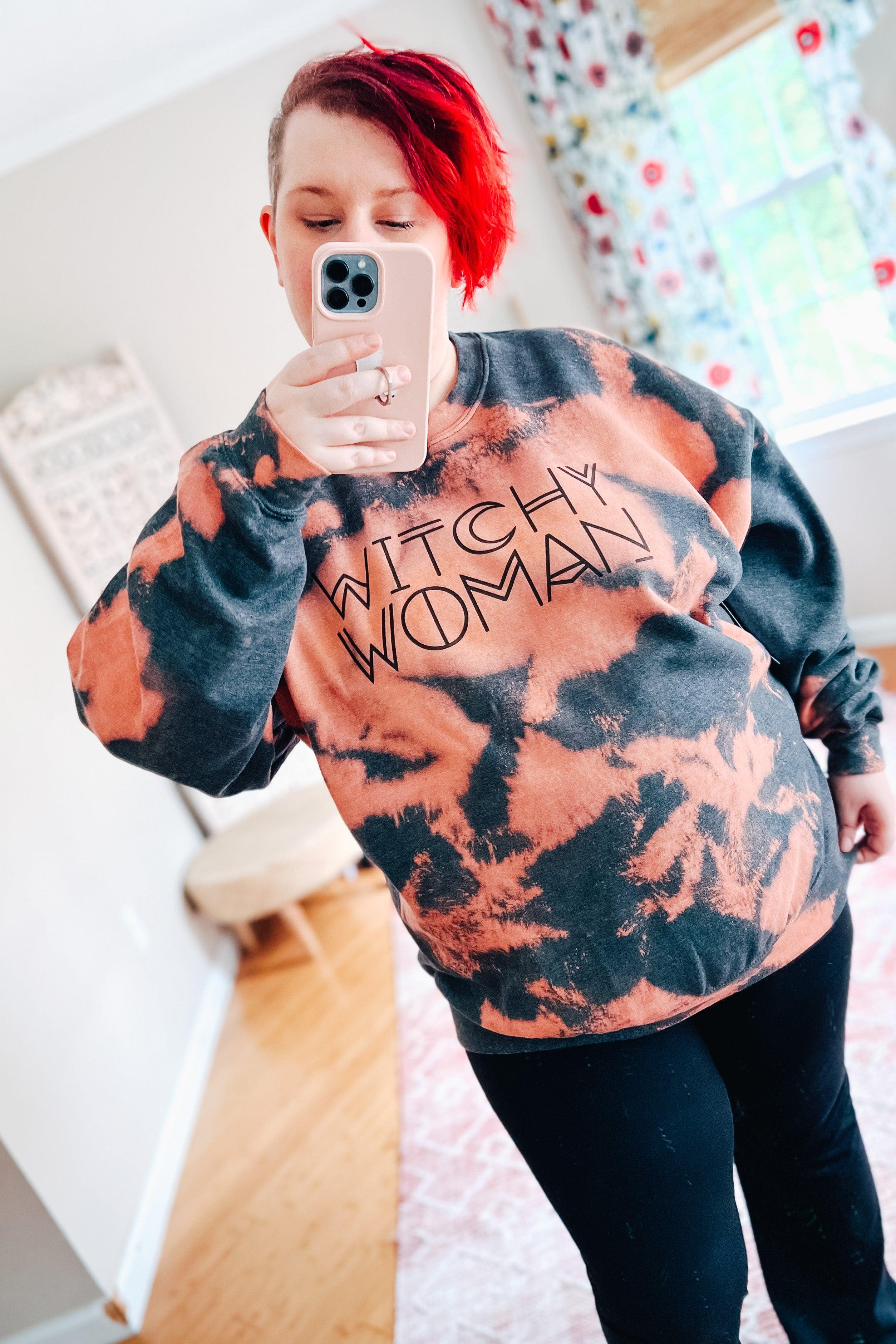 Woman with short red hair wearing bleach-dyed charcoal and pink sweatshirt with "Witchy Woman" text taking mirror selfie indoors