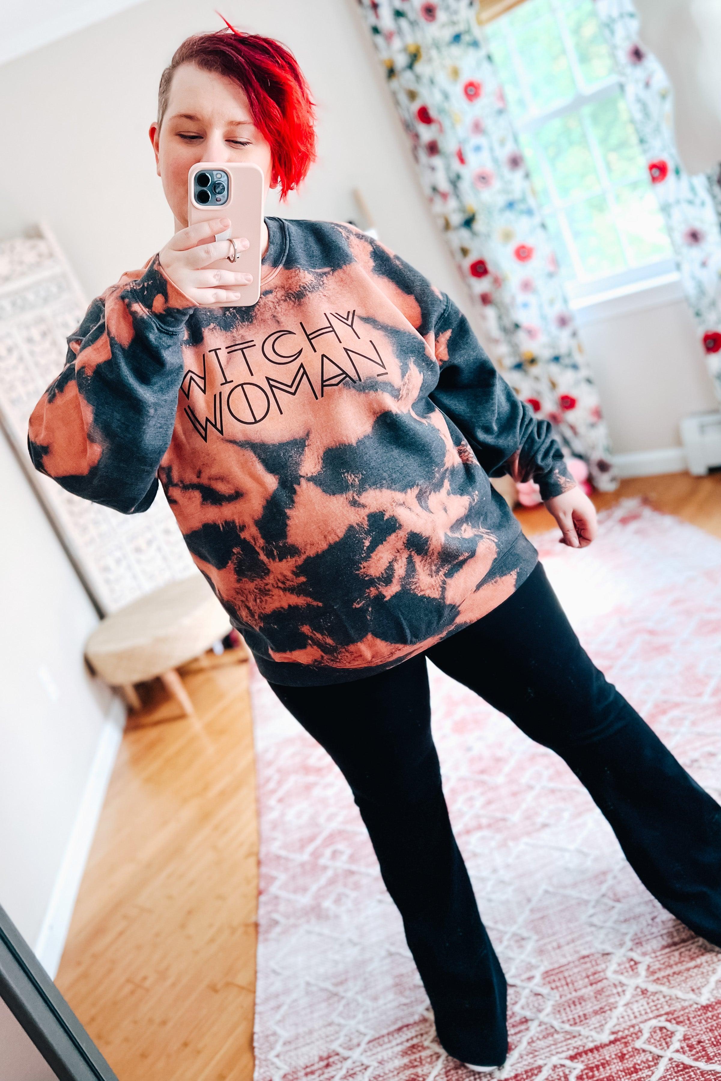 Woman with bright red hair wearing charcoal and pink bleached sweatshirt with "Witchy Woman" text in bright room