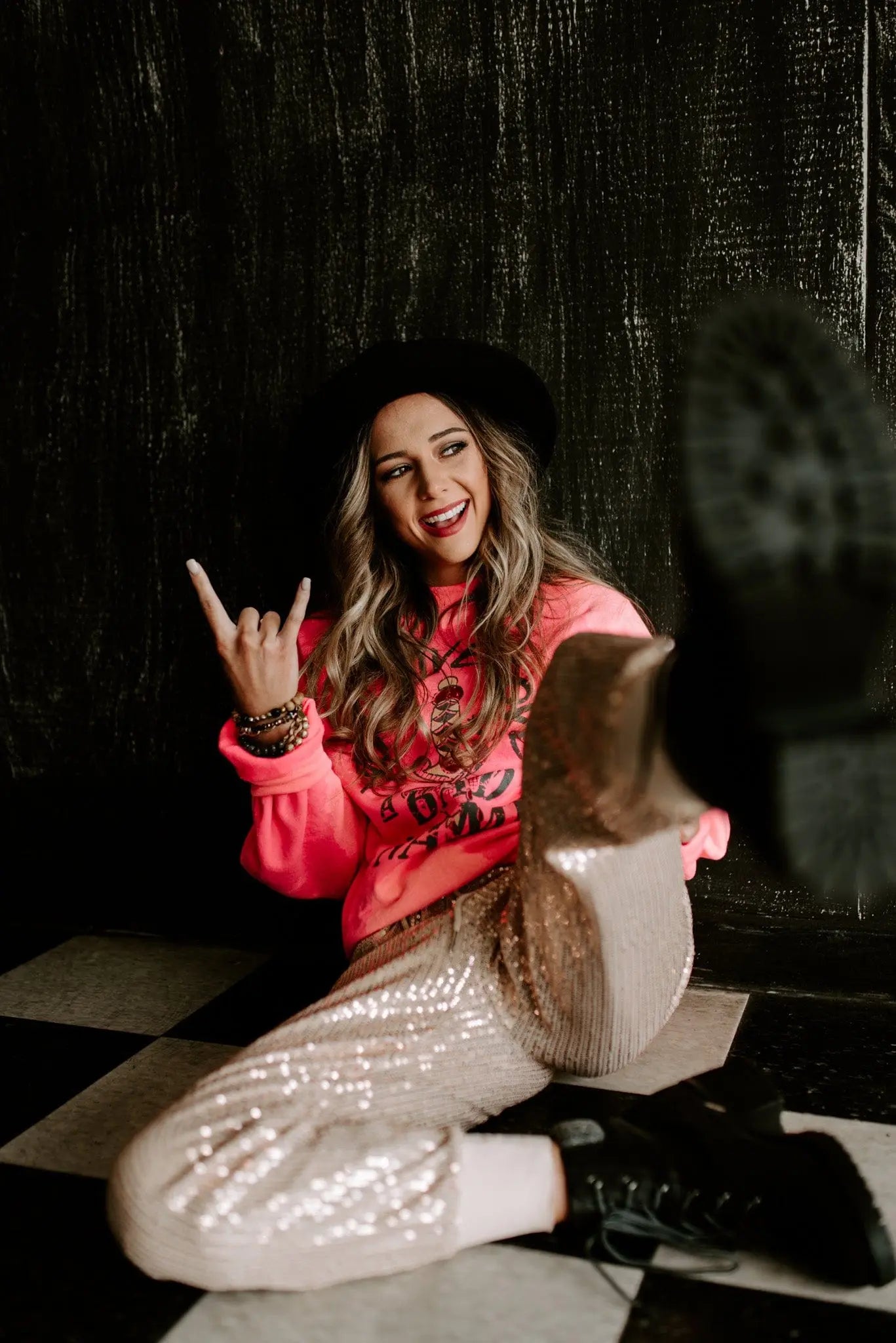 Woman in bright pink bleached pullover and sequined pants sitting on black and white floor with black hat and boots