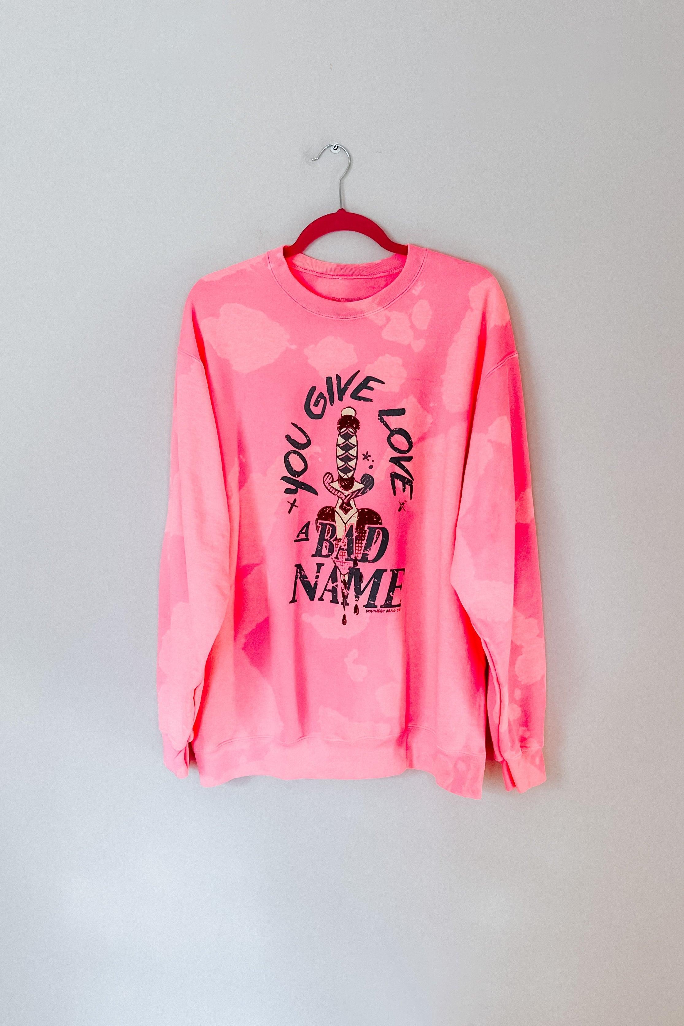 Pink bleached pullover sweatshirt with black graphic text and sword illustration hanging on red hanger