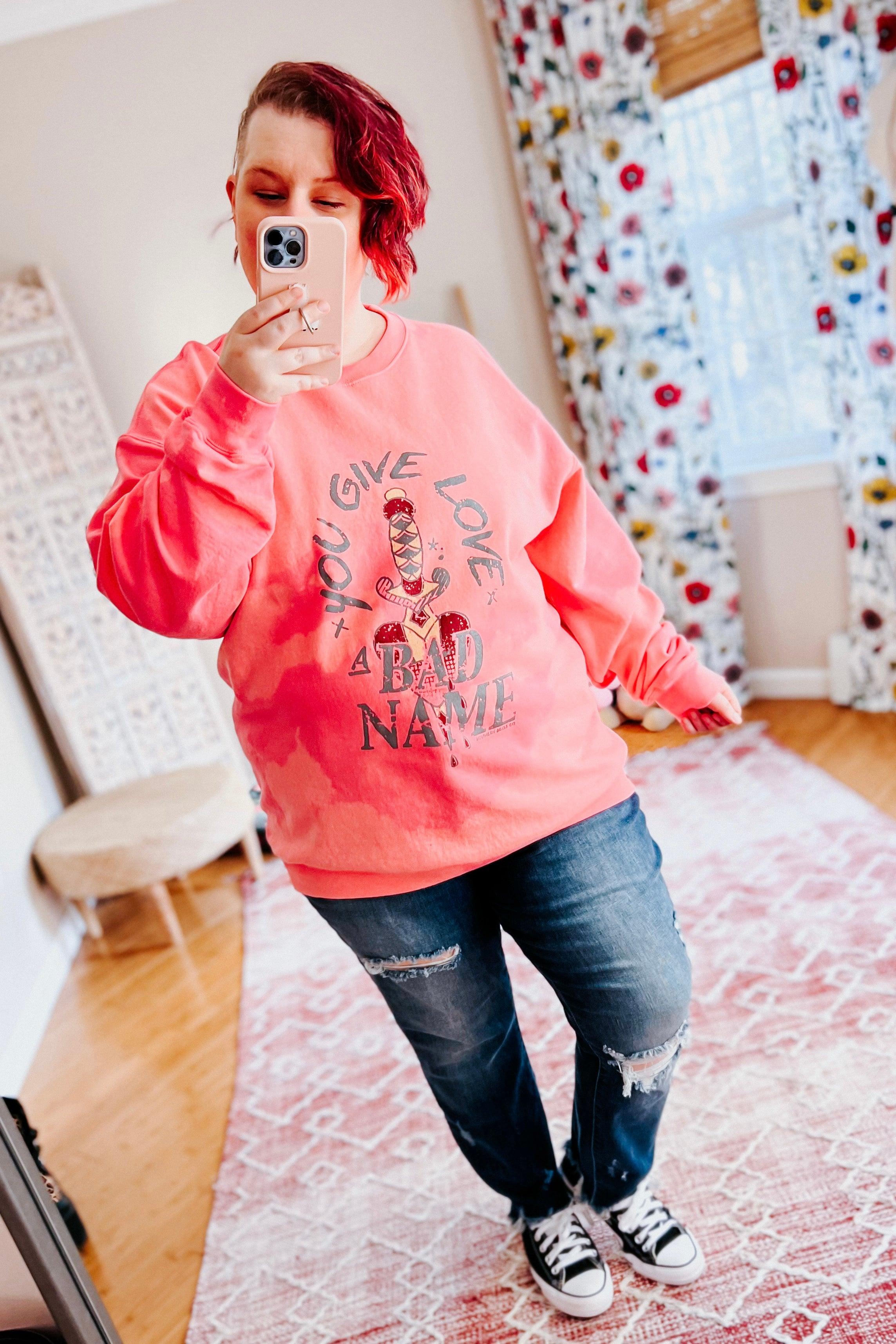 Woman with red hair wearing a pink bleached pullover with graphic text paired with ripped jeans and sneakers indoors