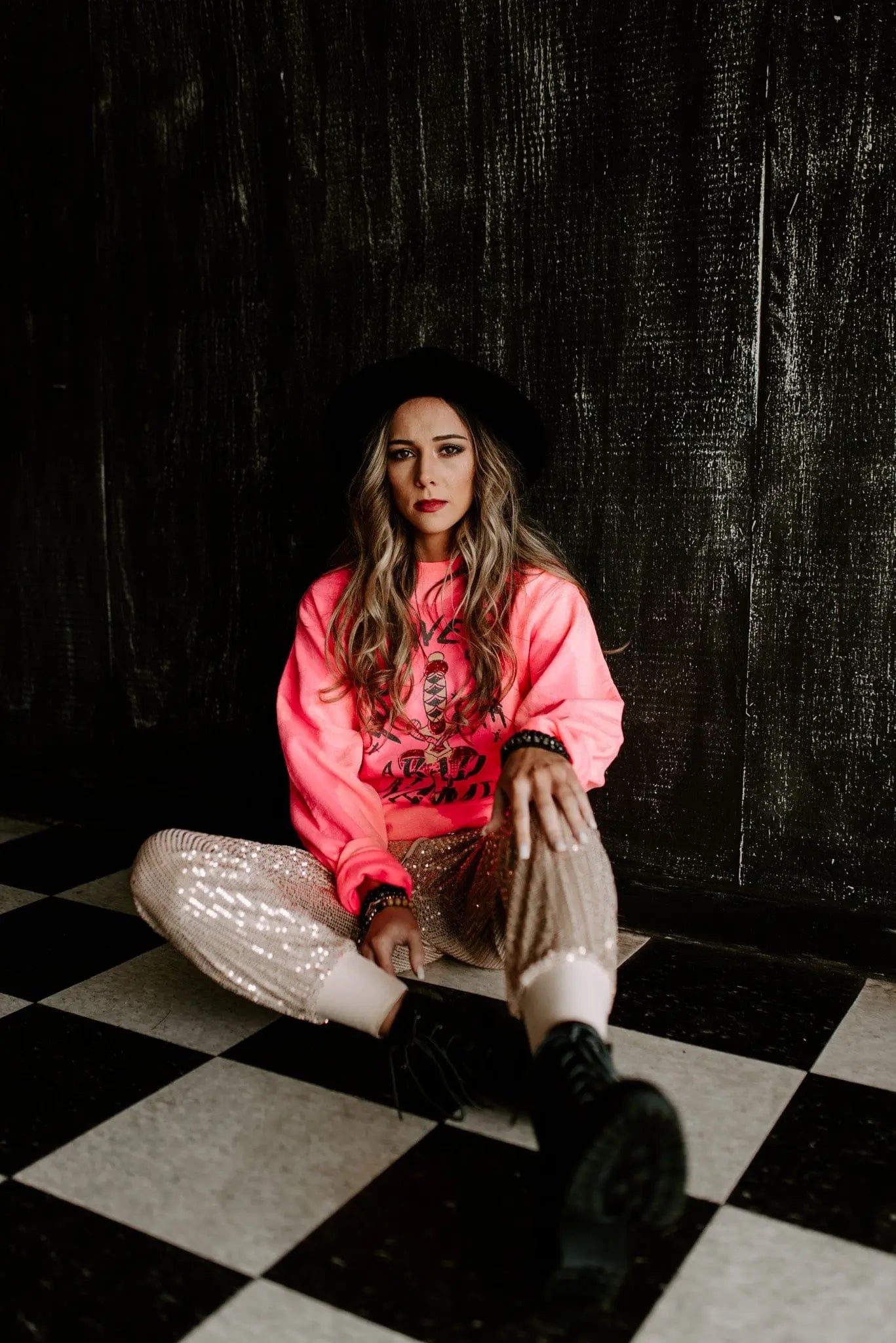 Woman wearing bright pink bleached pullover with graphic design paired with sequined pants and black boots seated on checkered floor