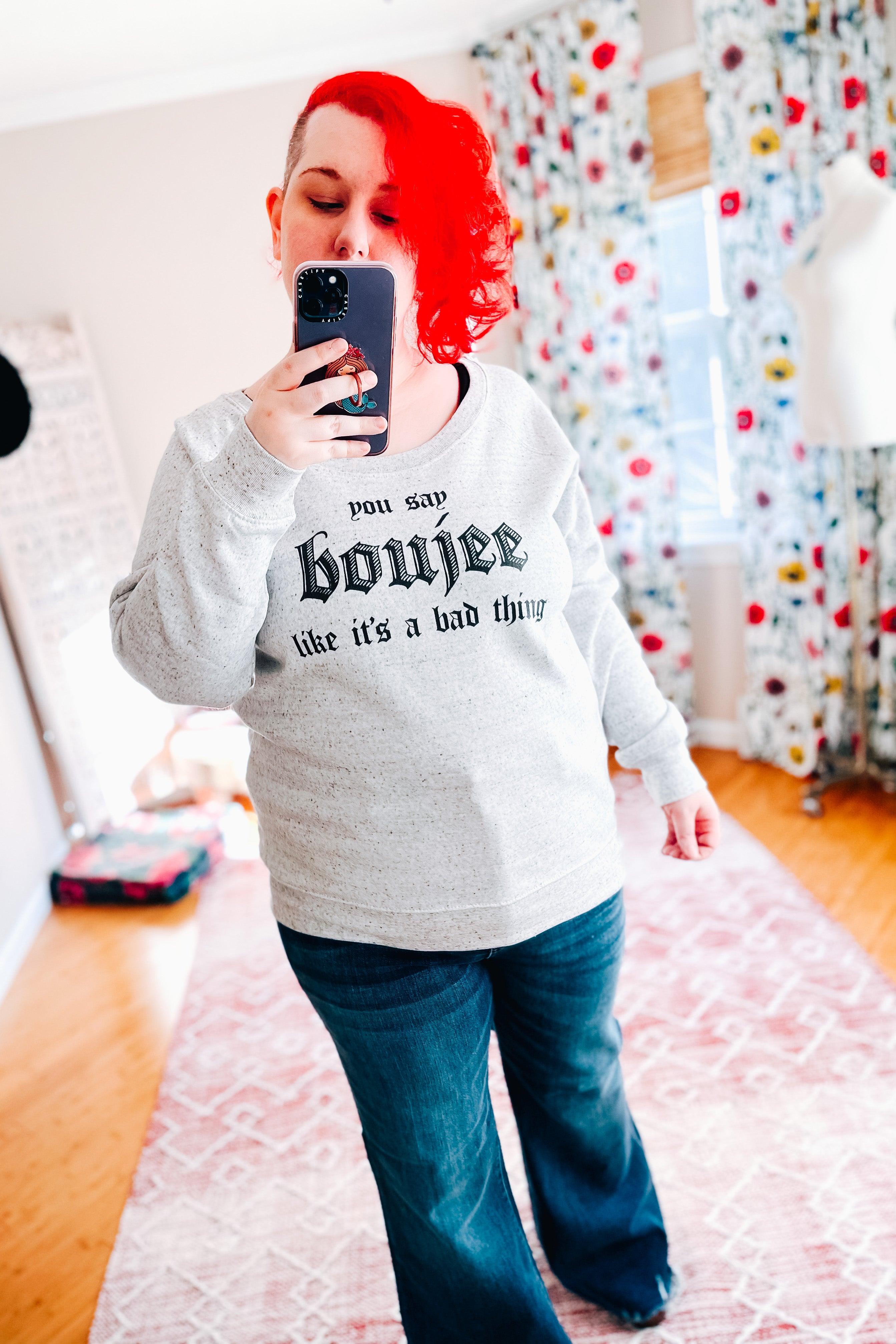 Woman with bright red hair wearing a grey speckled pullover with bold text paired with dark flared jeans indoors