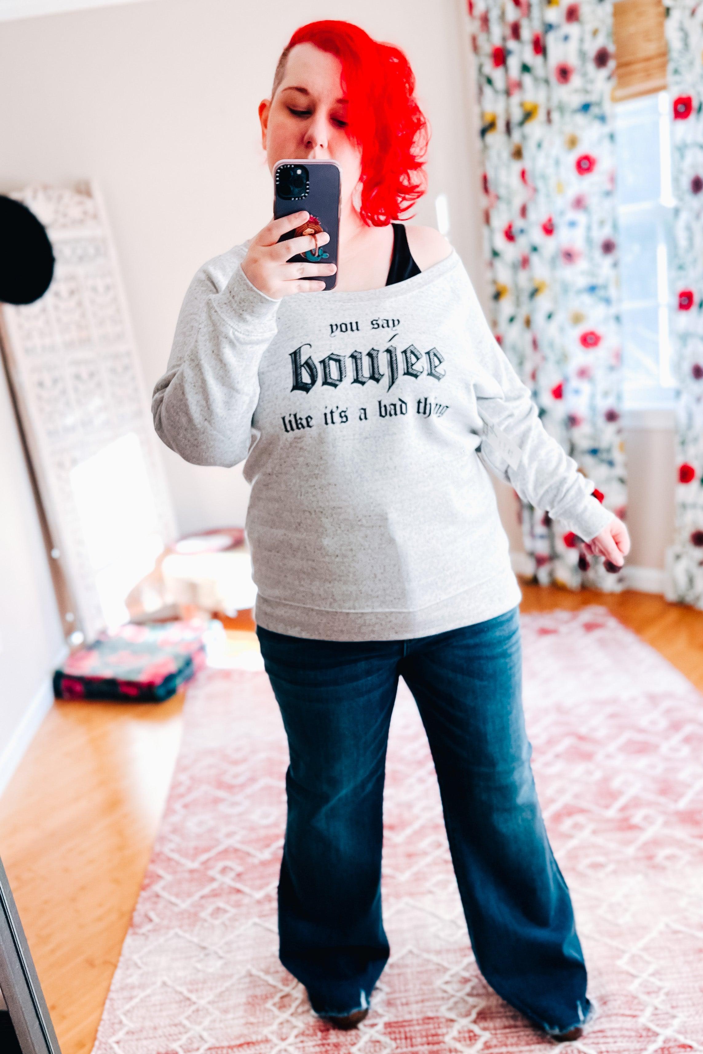 Woman with bright red hair wearing a grey speckled pullover with bold text paired with dark jeans indoors