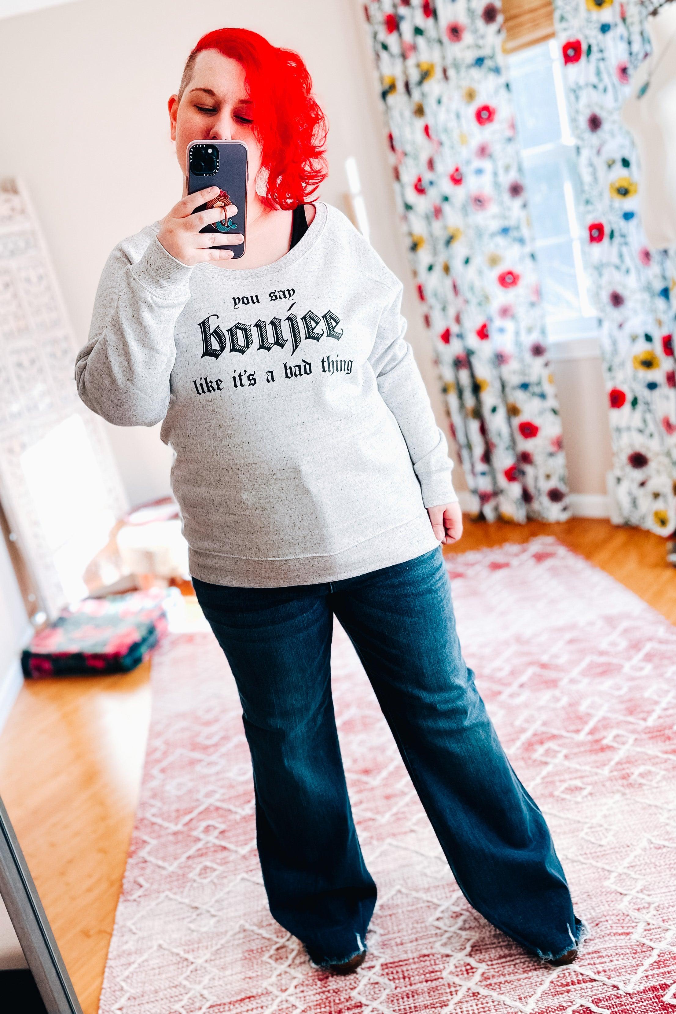 Woman with red asymmetrical hair wearing grey speckled pullover with bold text paired with dark wide-leg jeans indoors