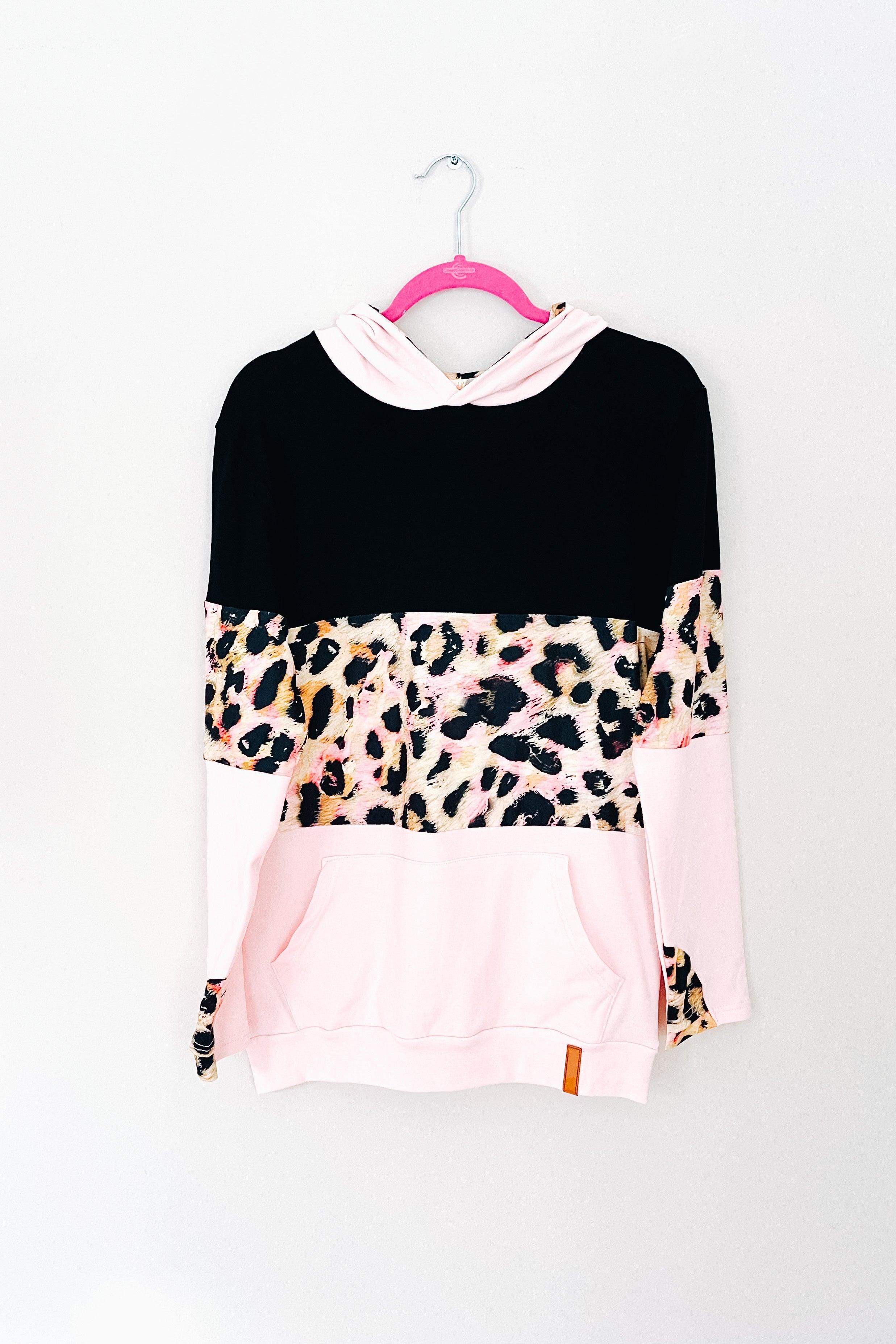 Hoodie with black upper, pink lower panels, and bold leopard print across midsection and sleeves on hanger against white wall