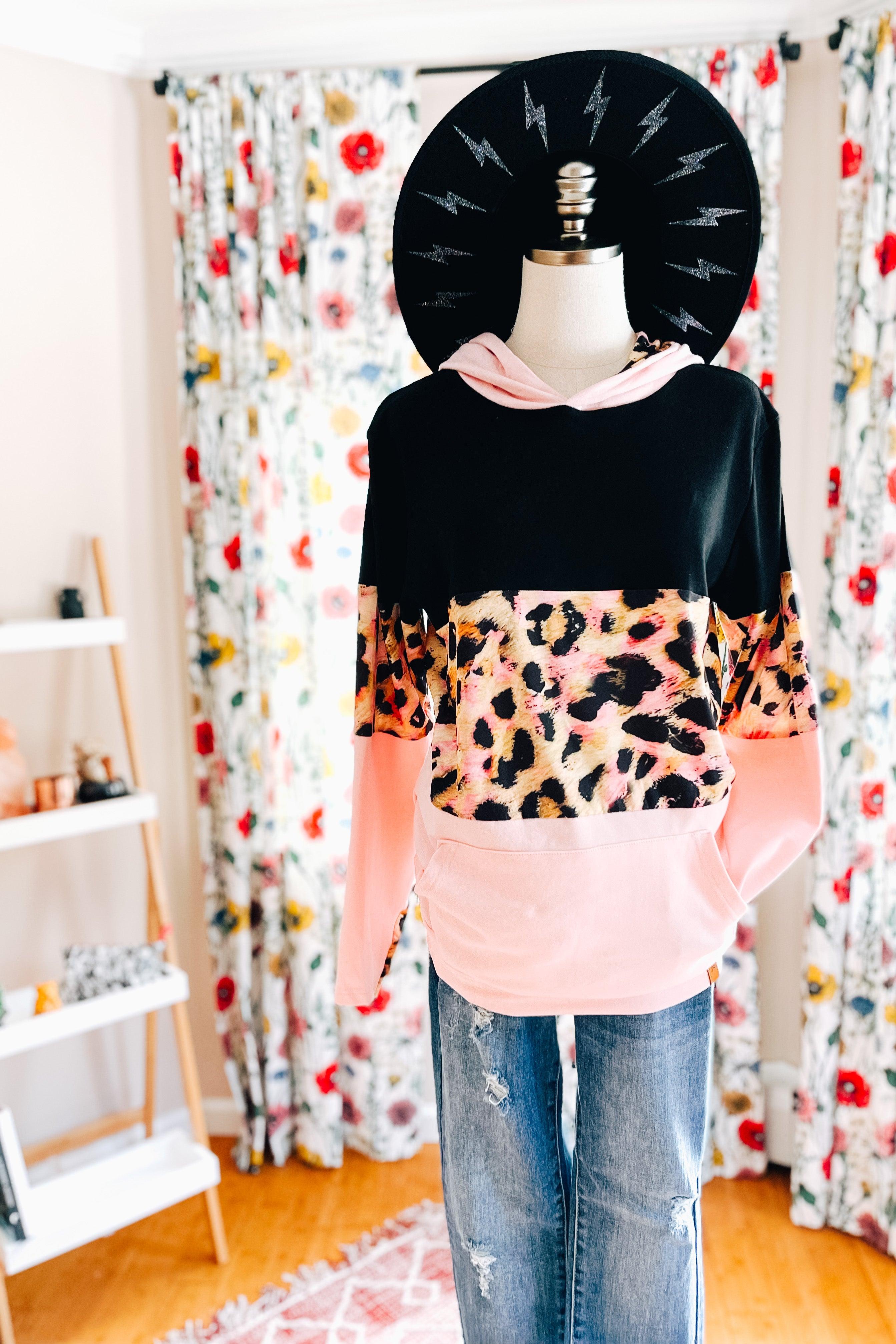 Mannequin dressed in black and pink hoodie with leopard print panel paired with distressed blue jeans and black hat indoors