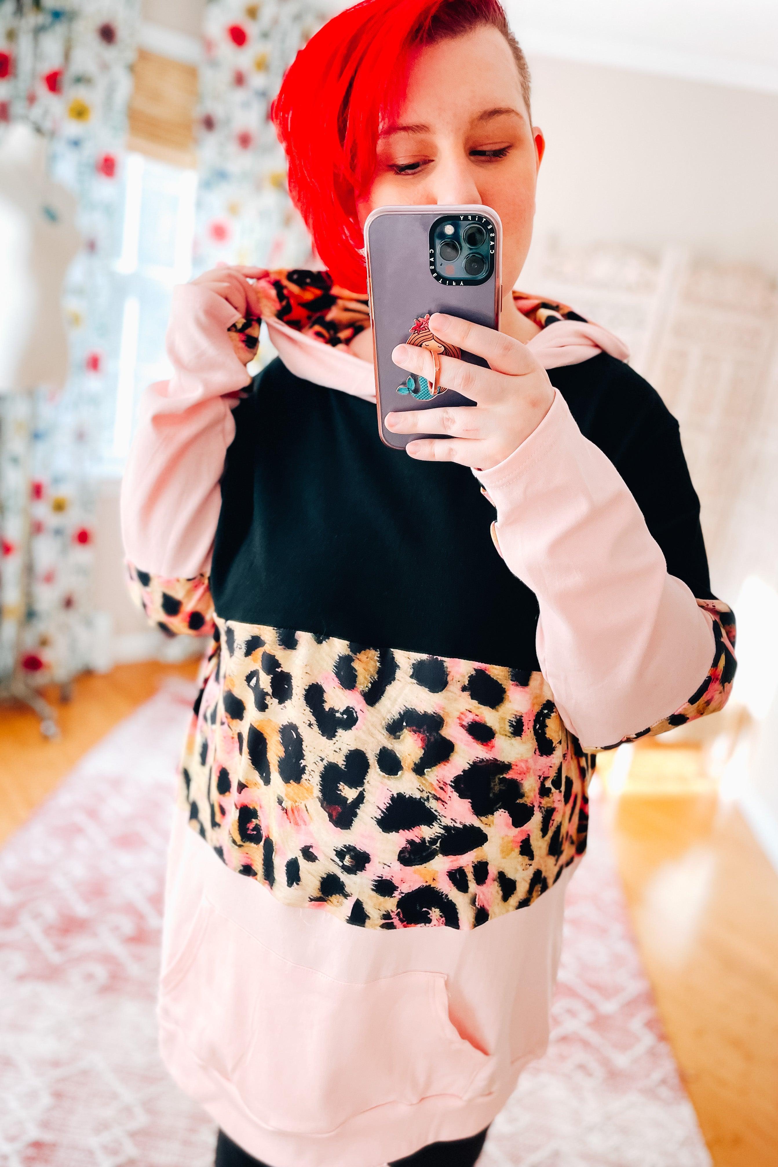 Woman wearing black and pink hoodie with leopard print panel taking indoor mirror selfie with red hair