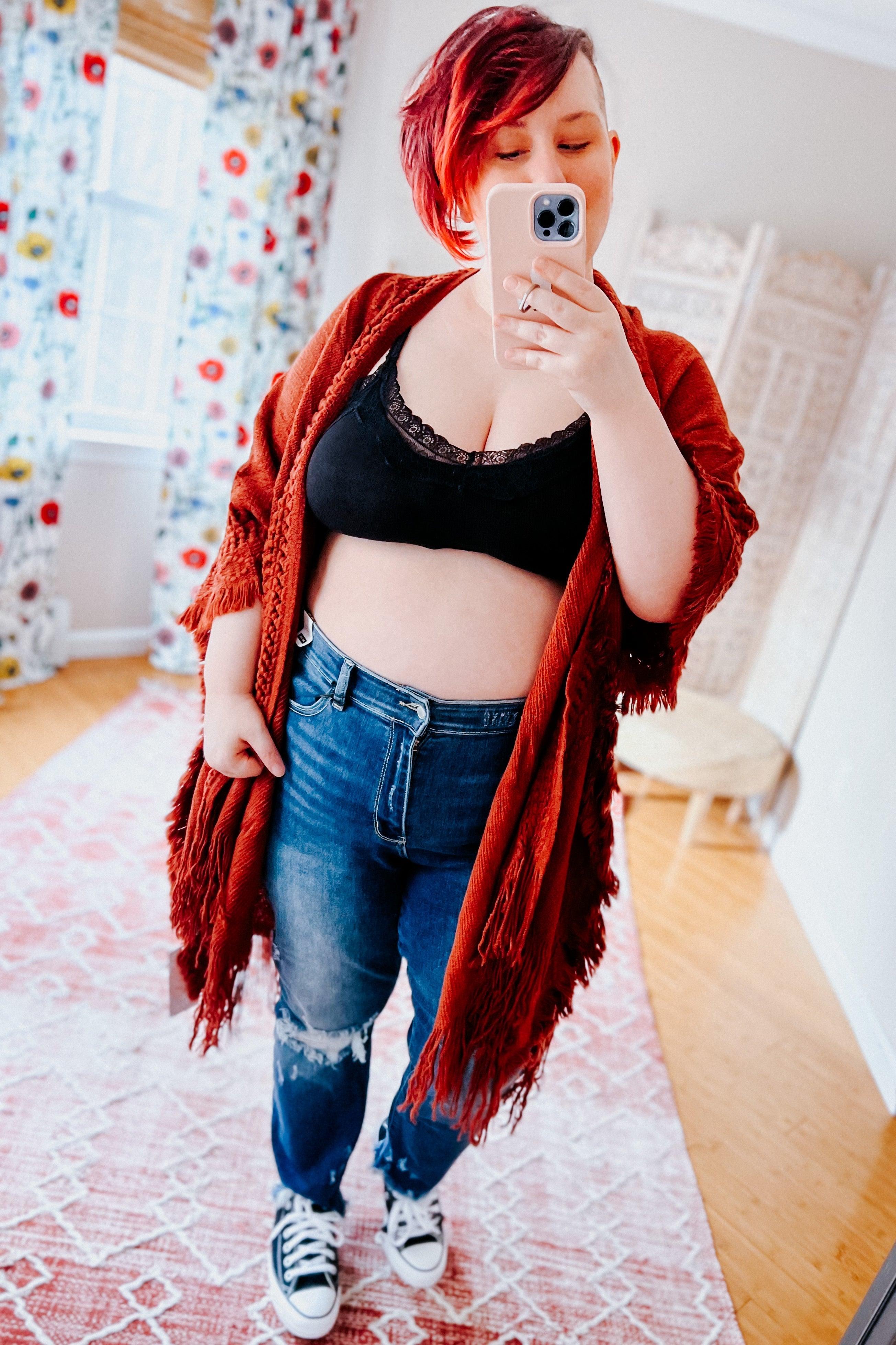 Woman with red hair wearing black lace trim bralette and rust fringed cardigan taking mirror selfie in cozy room