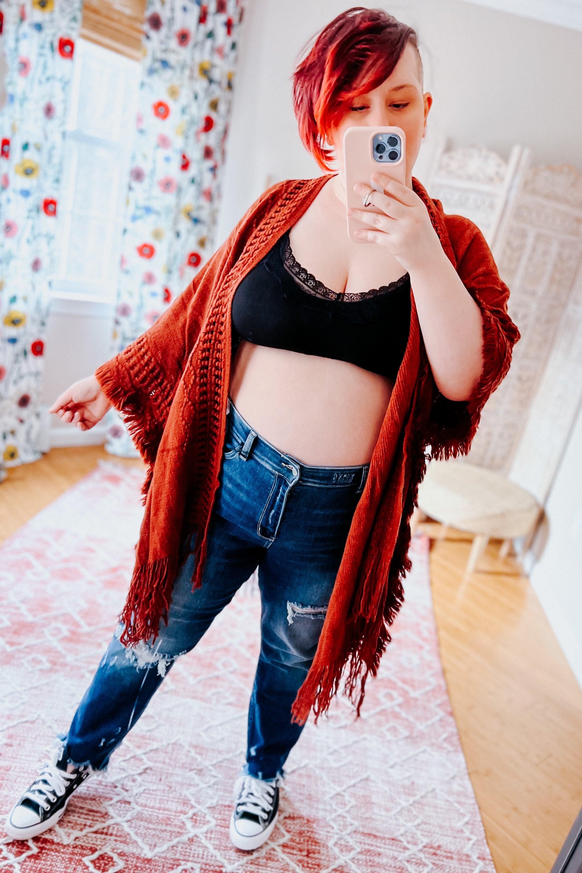 Woman with short red hair wearing black lace-trim bralette, rust fringe cardigan, distressed jeans, and sneakers taking selfie indoors