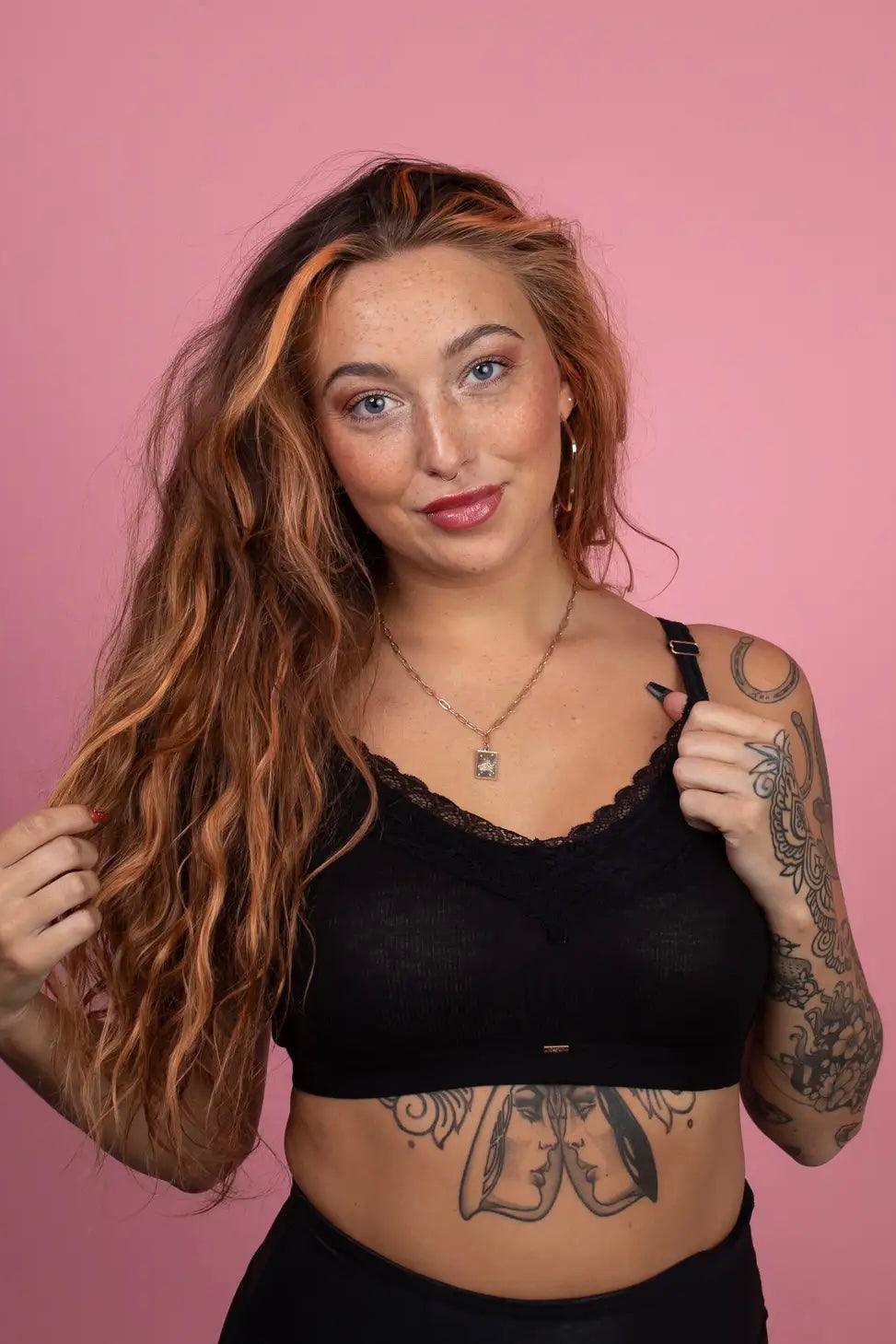 Woman with long wavy hair wearing black lace-trimmed bralette and tattoos on arm and stomach against pink backdrop