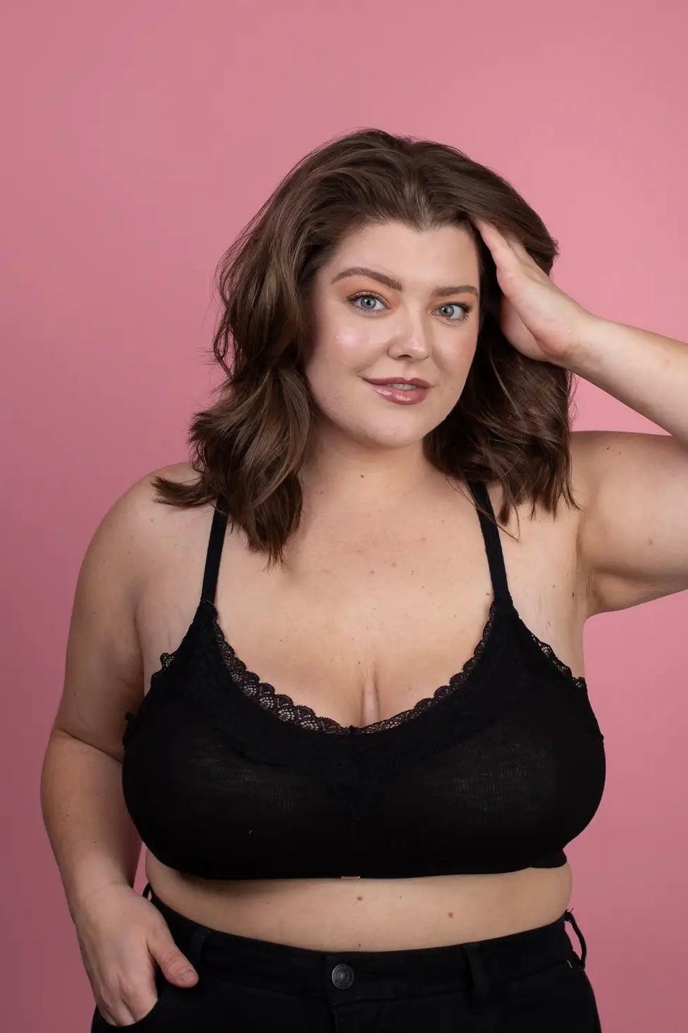 Smiling plus-size woman wearing black lace trim bralette with relaxed expression and hand in hair against pink background