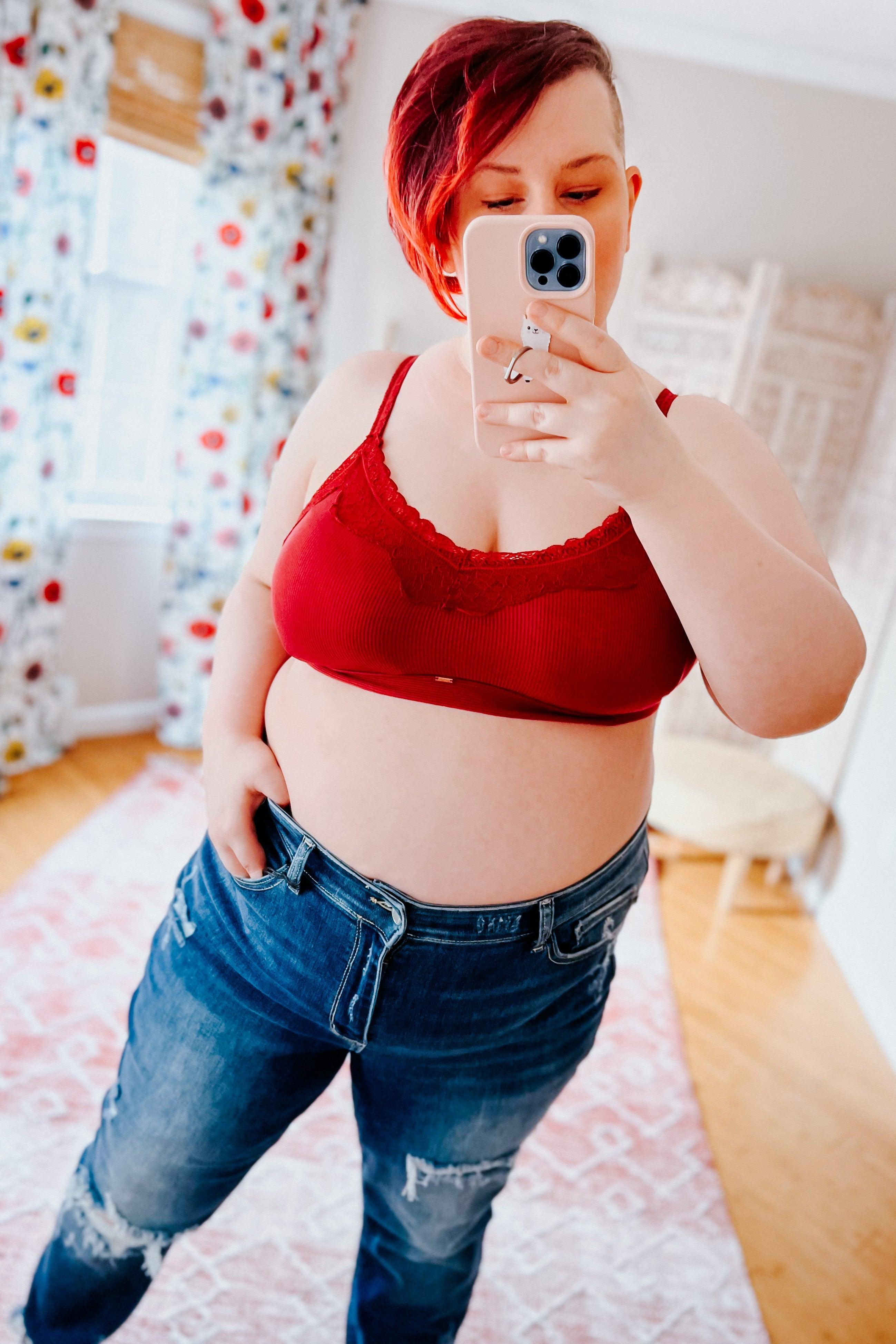 Plus-size woman with short red hair wearing wine lace trim bralette and distressed blue jeans taking mirror selfie indoors