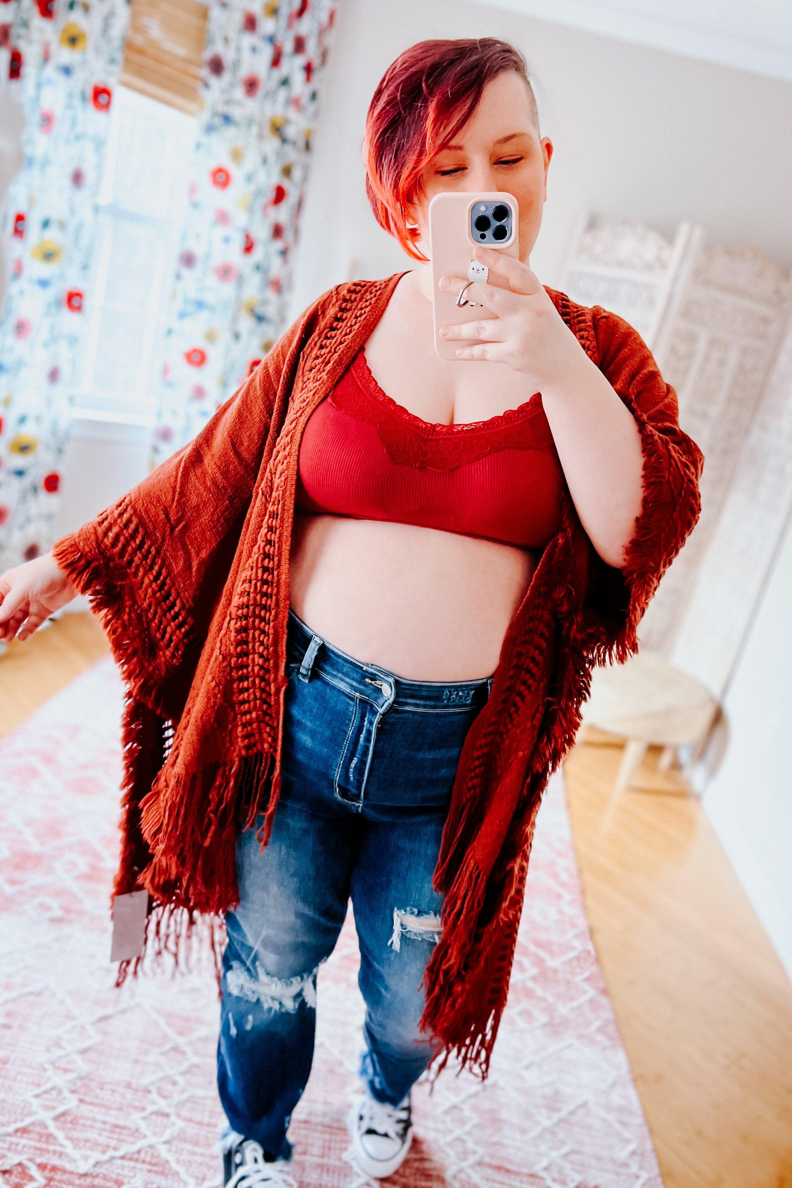Woman with short red hair wearing wine-colored lace trim bralette and distressed jeans taking selfie in bright room with floral curtains