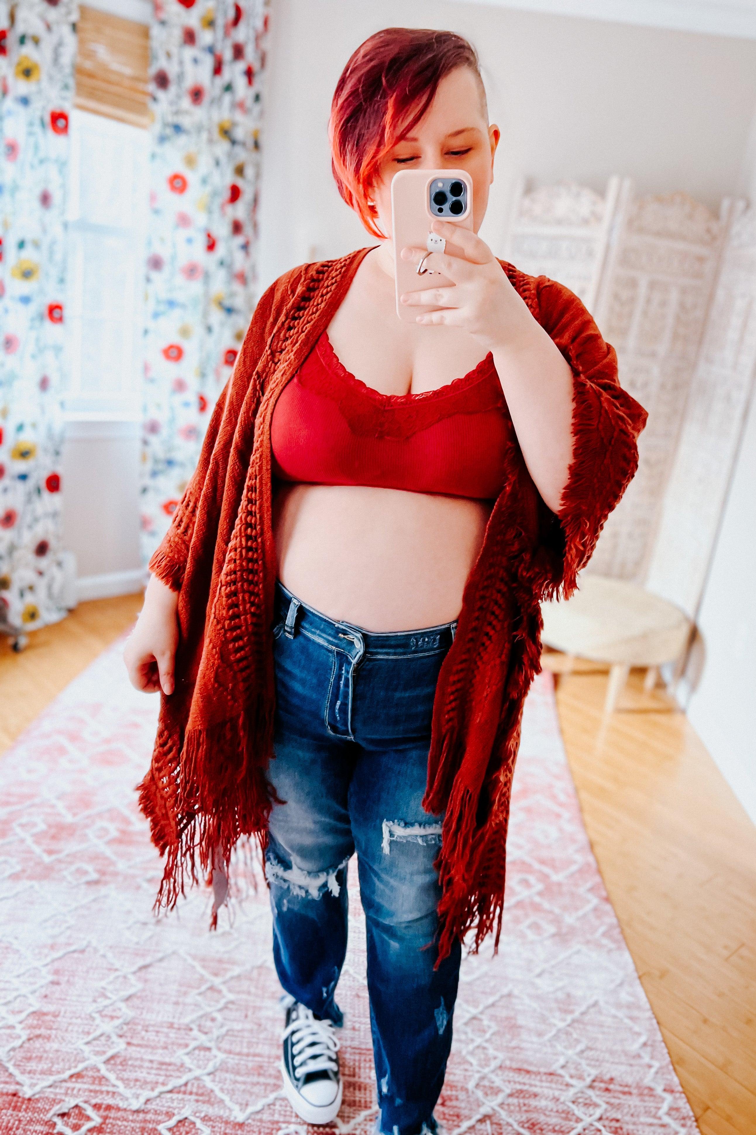 Plus-size woman with short red hair wearing wine lace-trim bralette under rust fringe cardigan and distressed jeans taking selfie