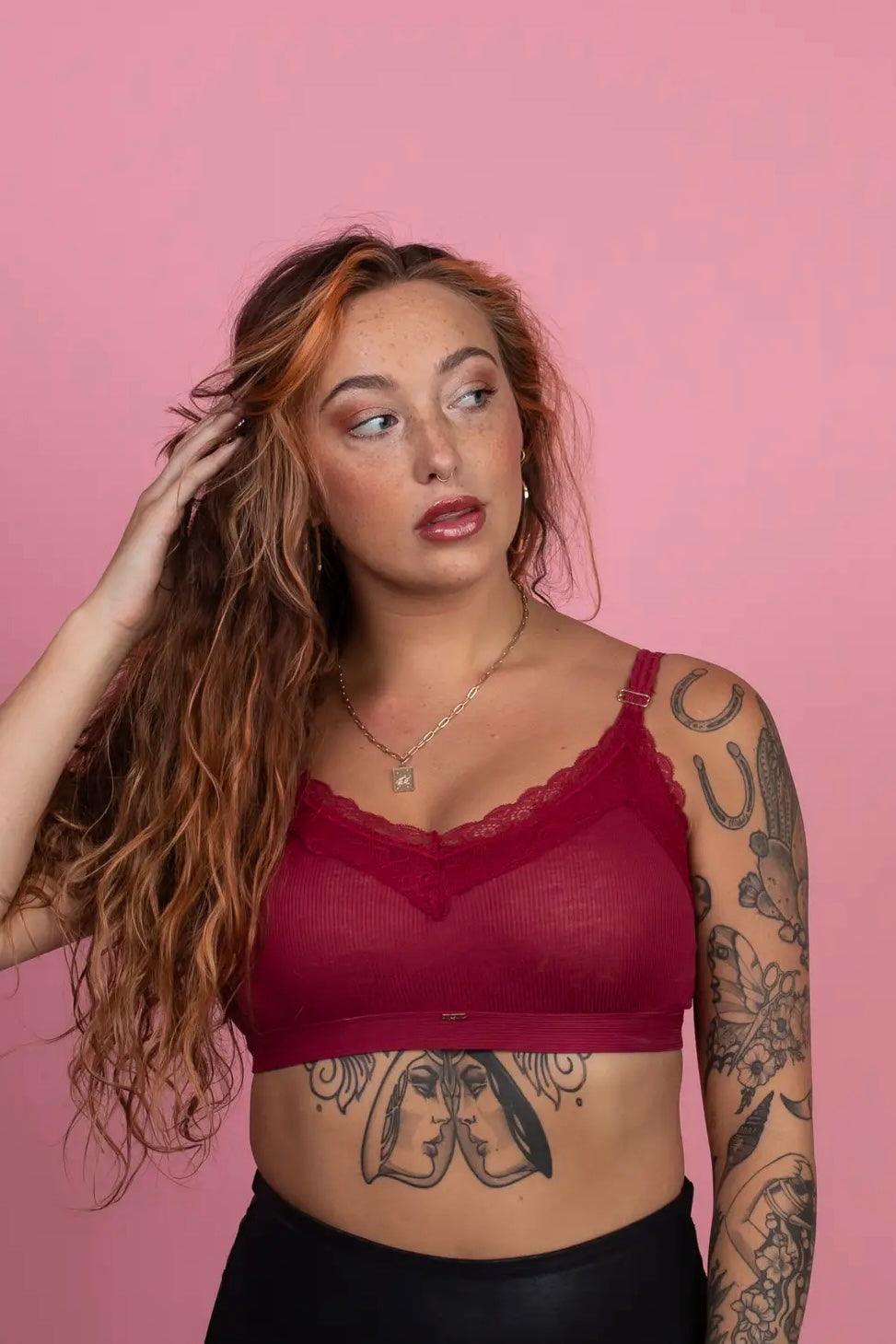 Woman with long red hair and arm tattoos wearing wine-colored lace-trim bralette against pink backdrop