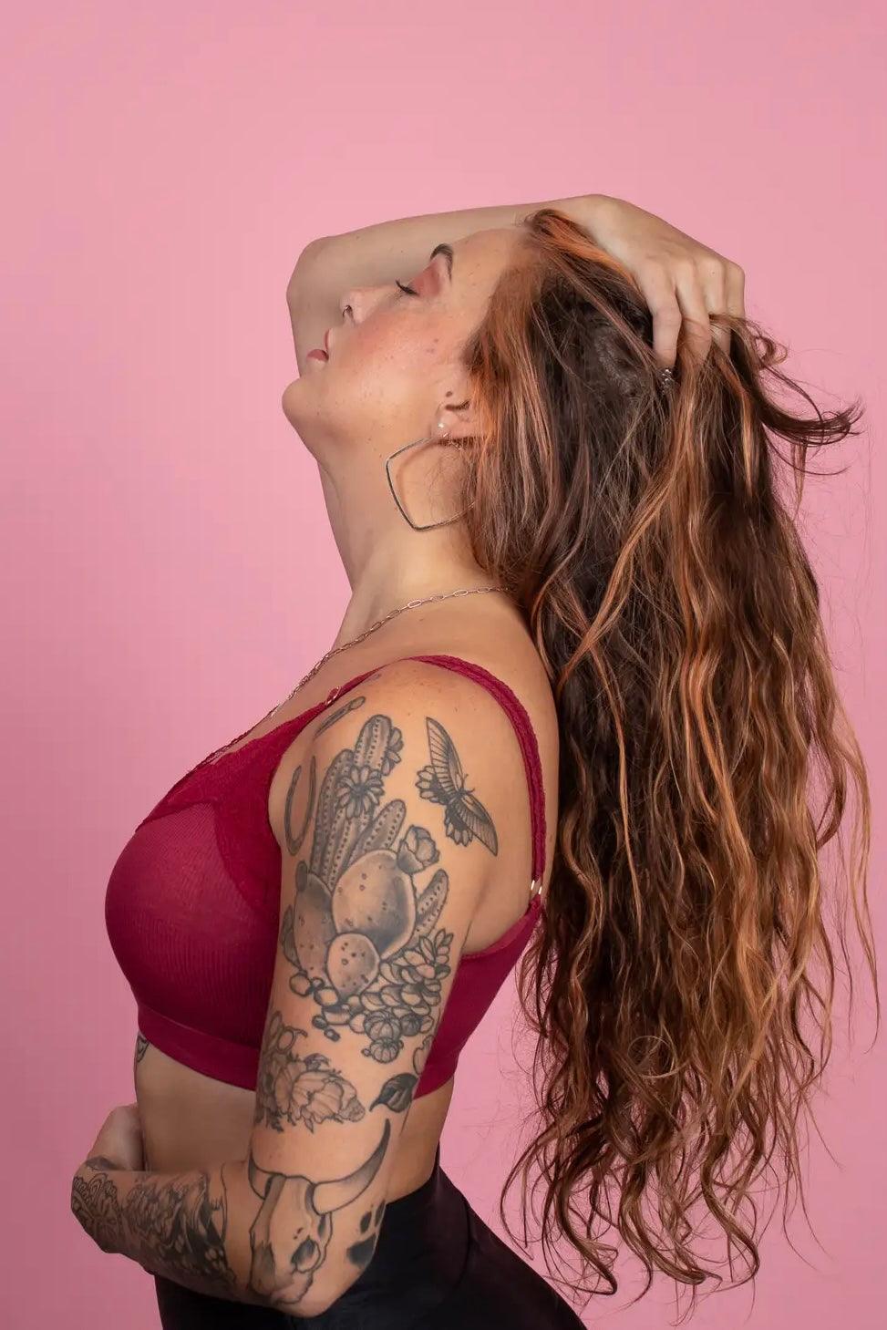 Woman with long wavy hair wearing wine-colored lace bralette and hoop earrings posing against pink background