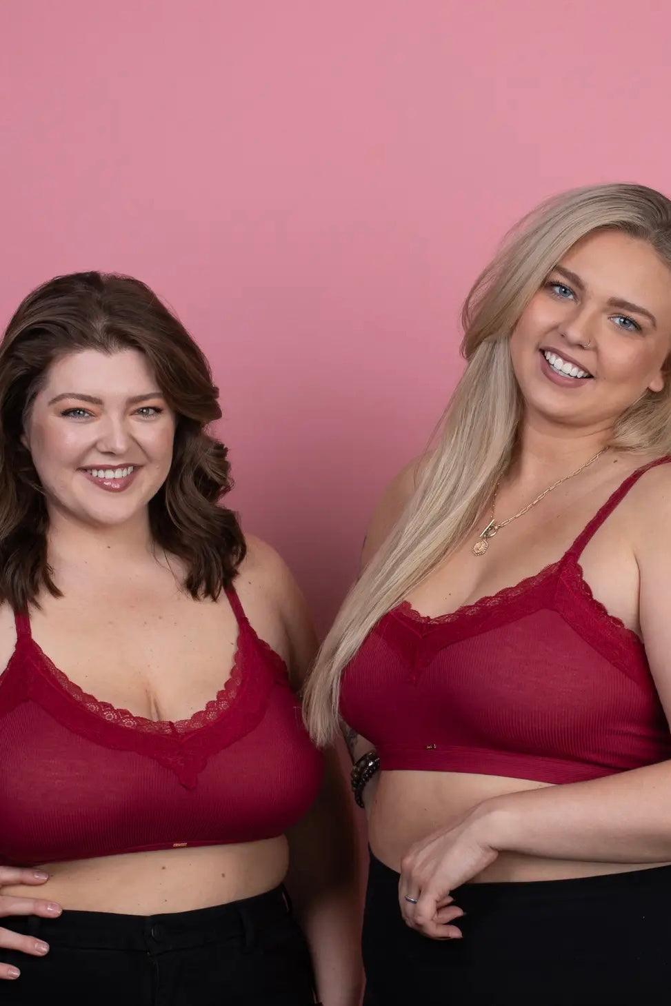 Two plus-size women smiling wearing wine red lace-trim bralettes styled with black bottoms against pink backdrop