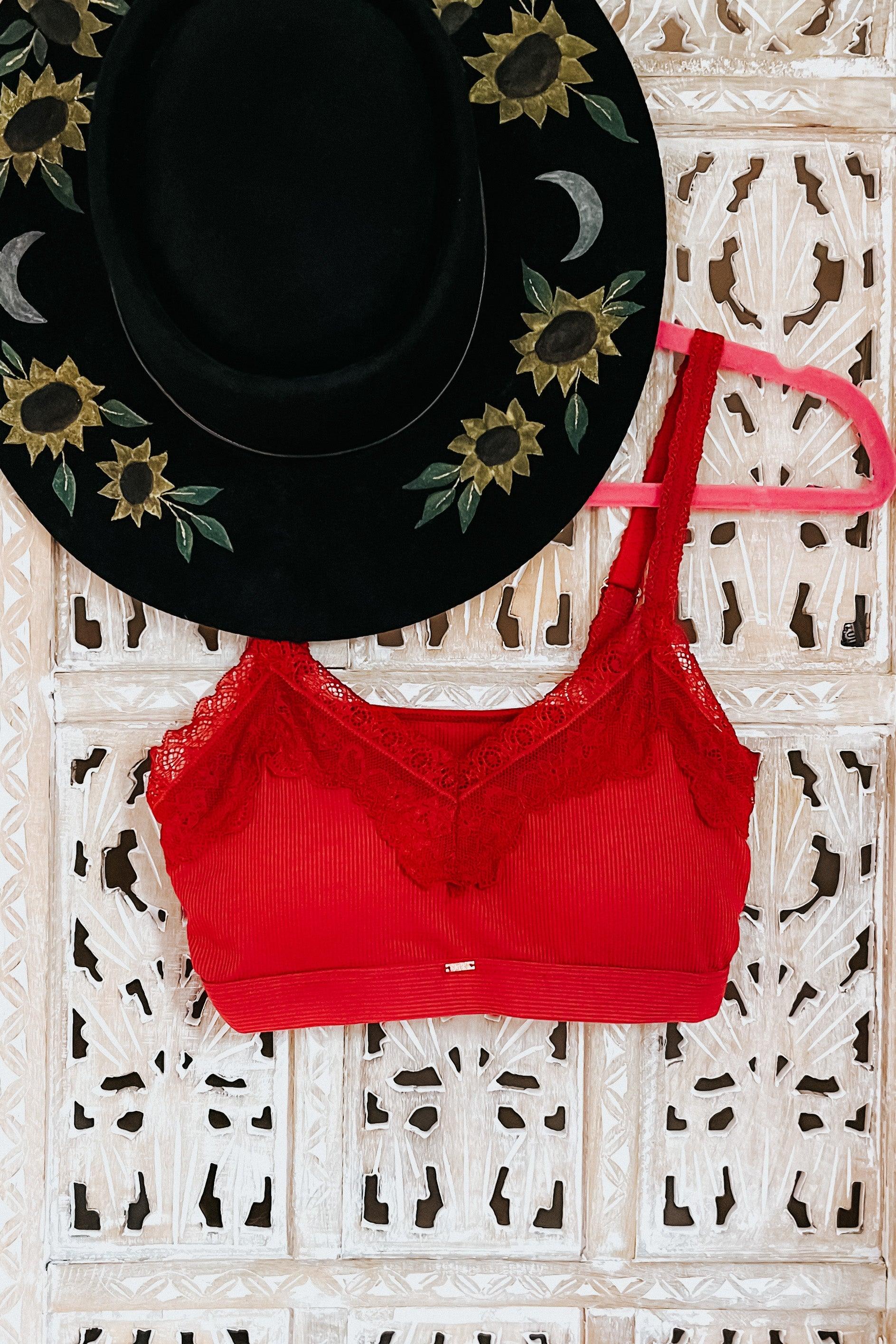 Wine-colored lace-trim bralette with ribbed fabric hanging on pink hanger beside black hat with sunflower and moon embroidery