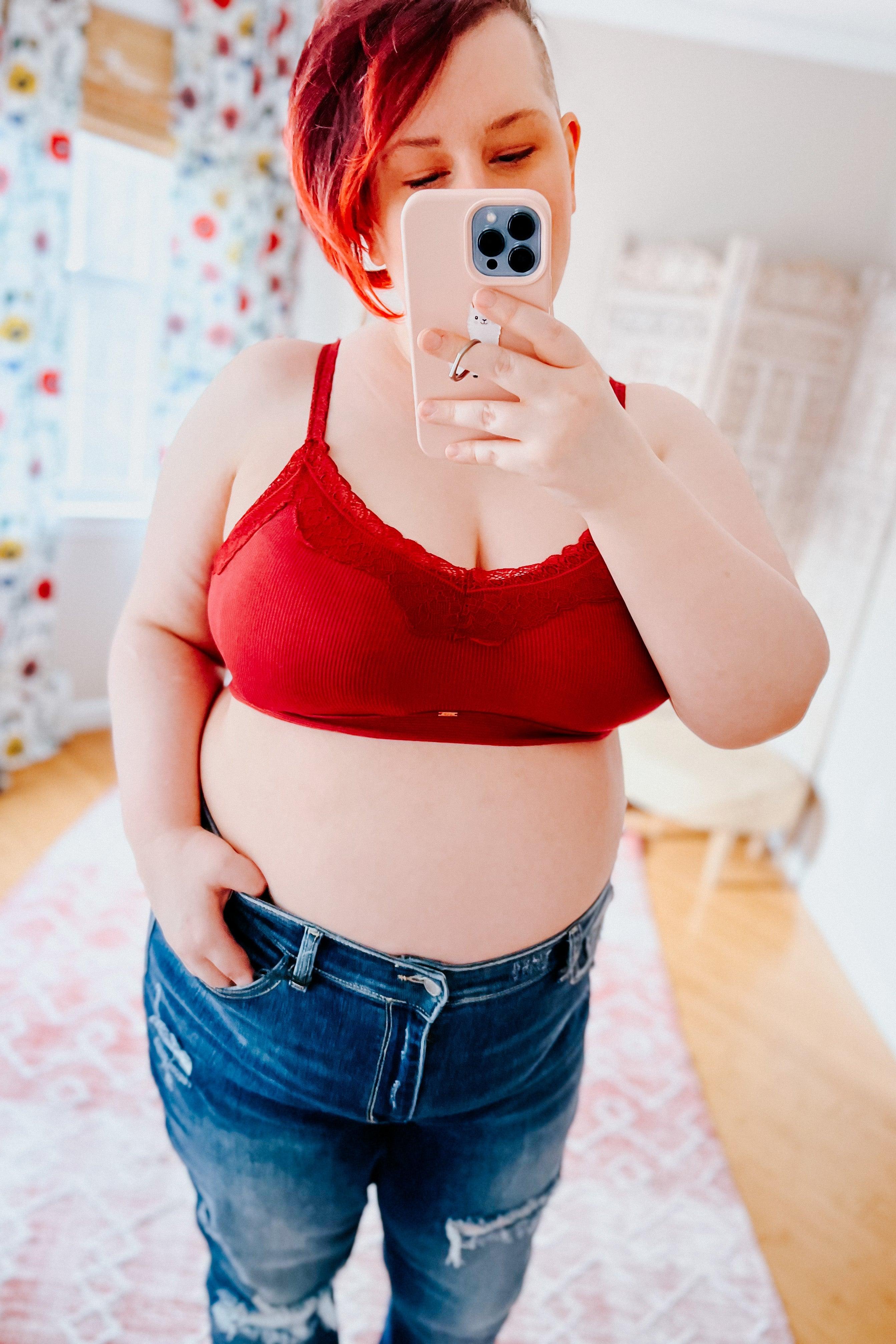Plus-size woman with short red hair wearing wine-colored lace trim bralette and distressed blue jeans, taking mirror selfie